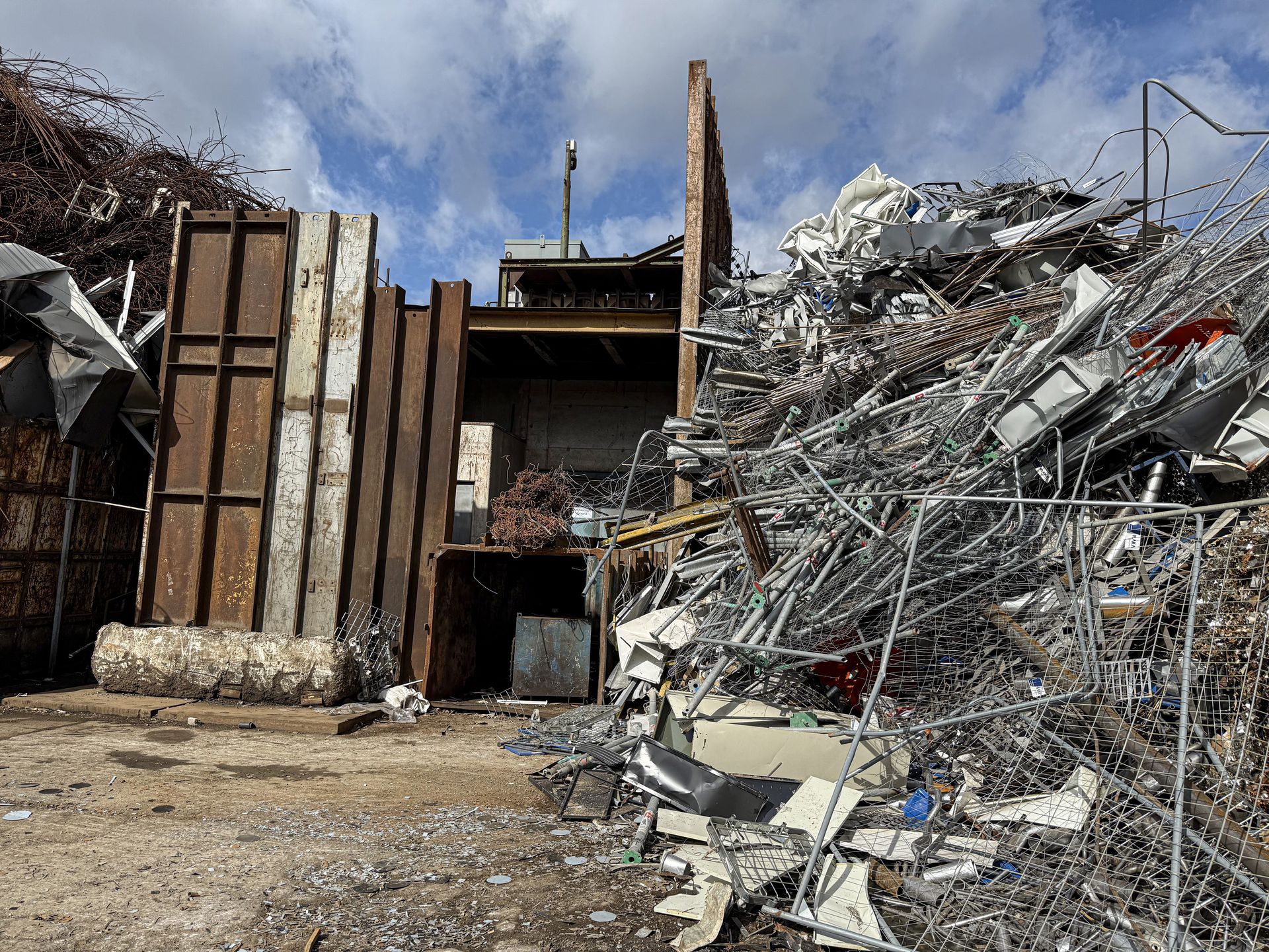 Scrap metal yard: piles of metal, large metal structures, and a small building under a cloudy sky.