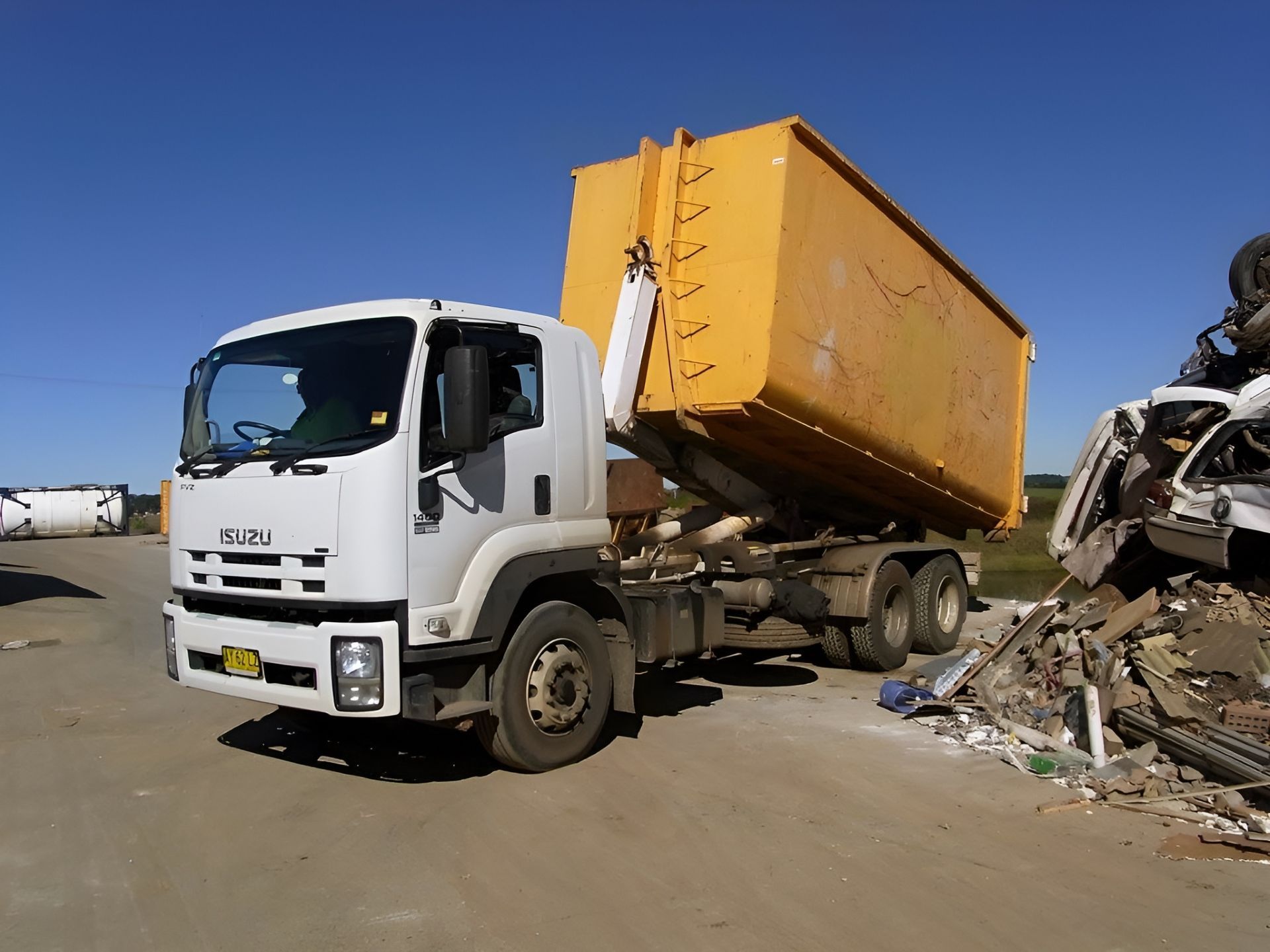 White Isuzu truck with yellow dumpster tilting, unloading waste at a recycling center under a blue sky.