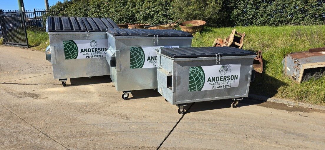 Three metal dumpsters on wheels, lined up on a gray asphalt surface next to green grass.