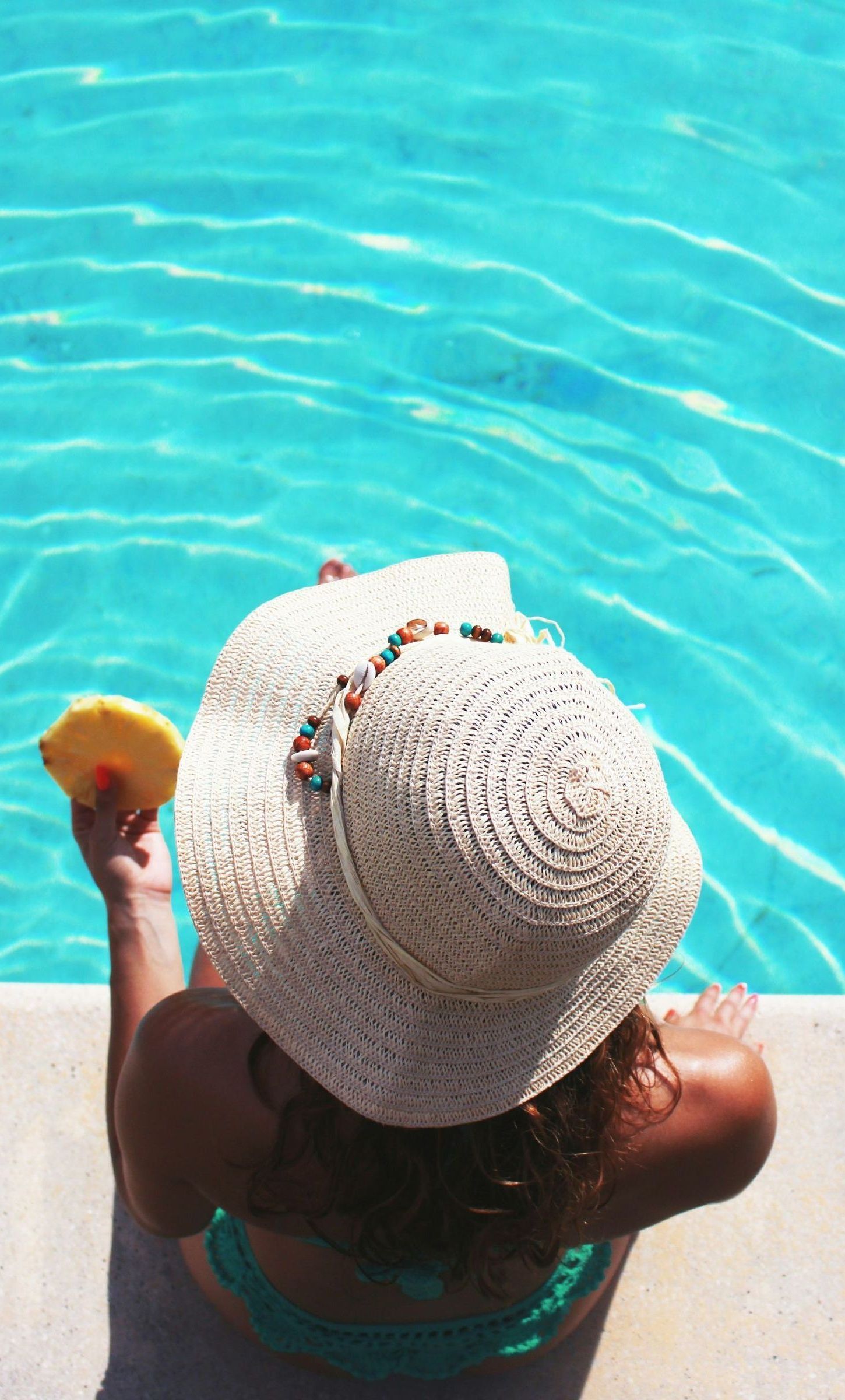 A woman is sitting on the edge of a swimming pool holding a pineapple slice.