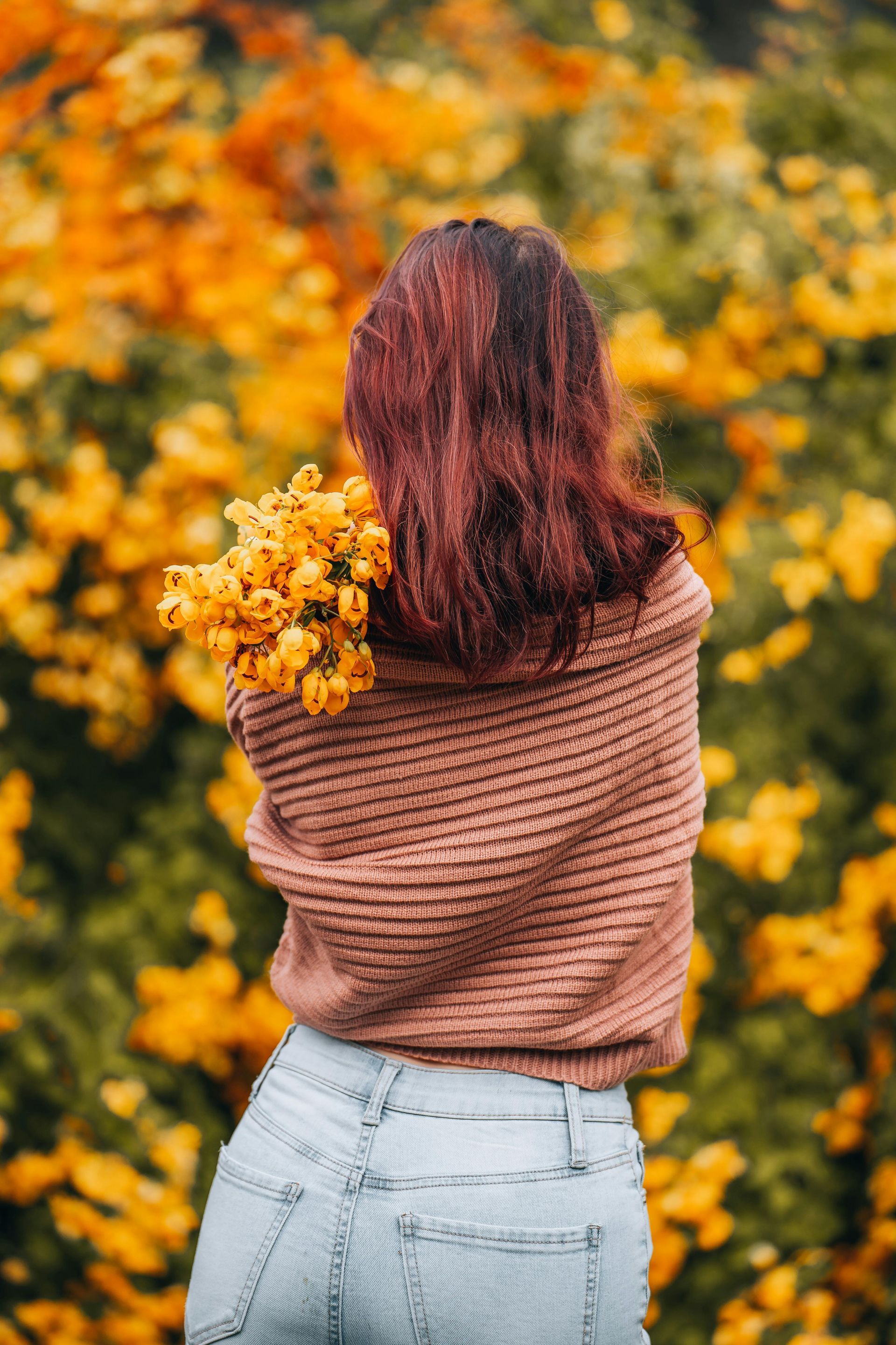 Vrouw in een bruine trui met gele bloemen in haar handen, staand voor gele bloesem.
