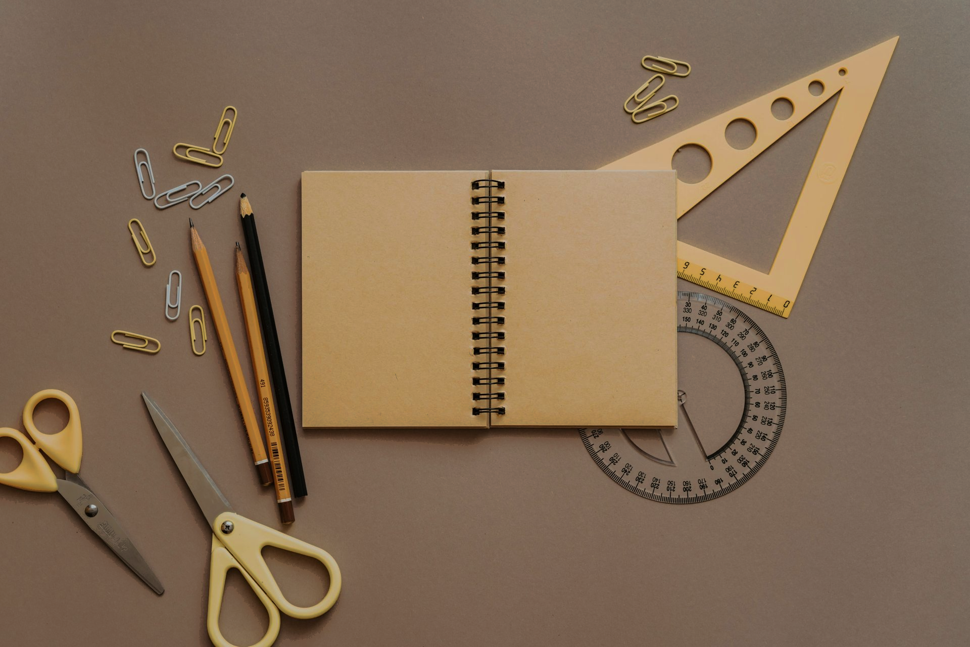 Open notitieboek, potloden, schaar en geometrische gereedschappen op een bruine ondergrond.