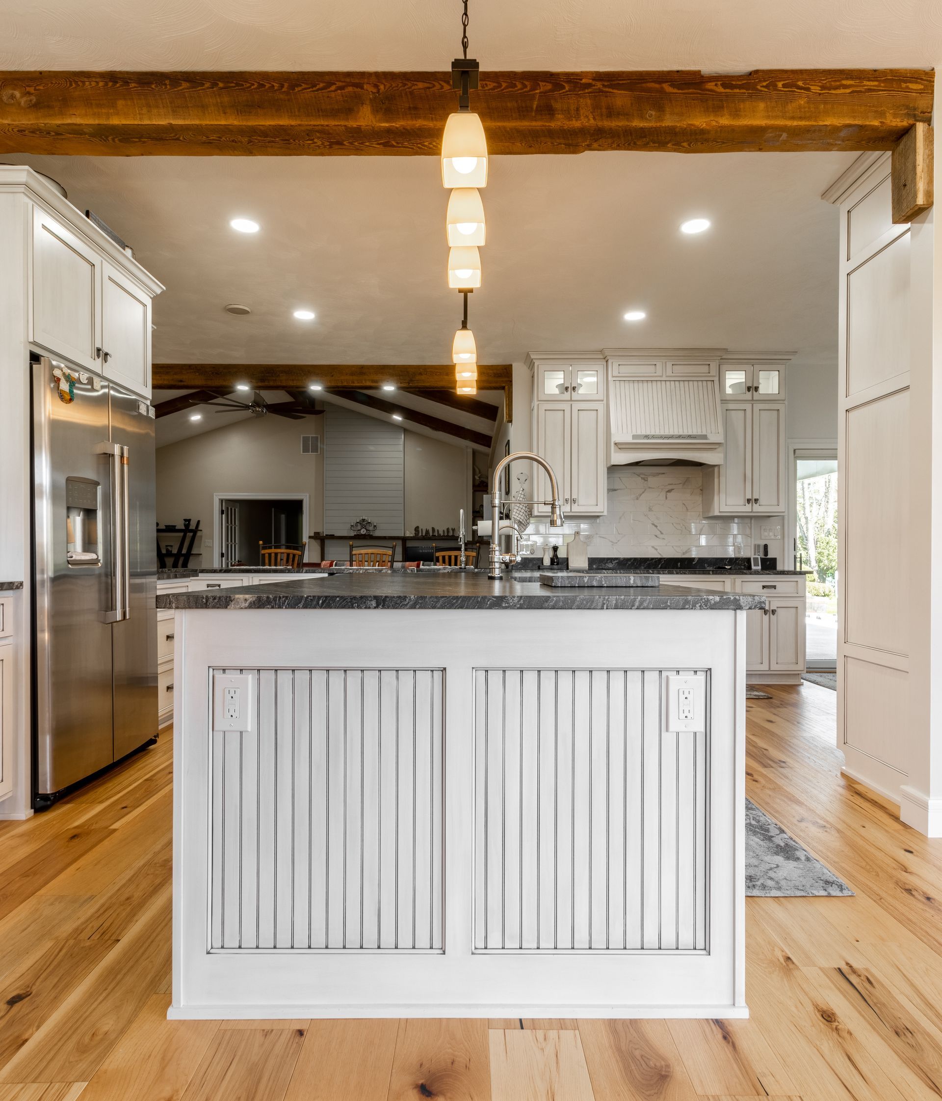 Kitchen with white cabinets, wood floors, and island. Stainless steel refrigerator, wooden beams, pendant lights.