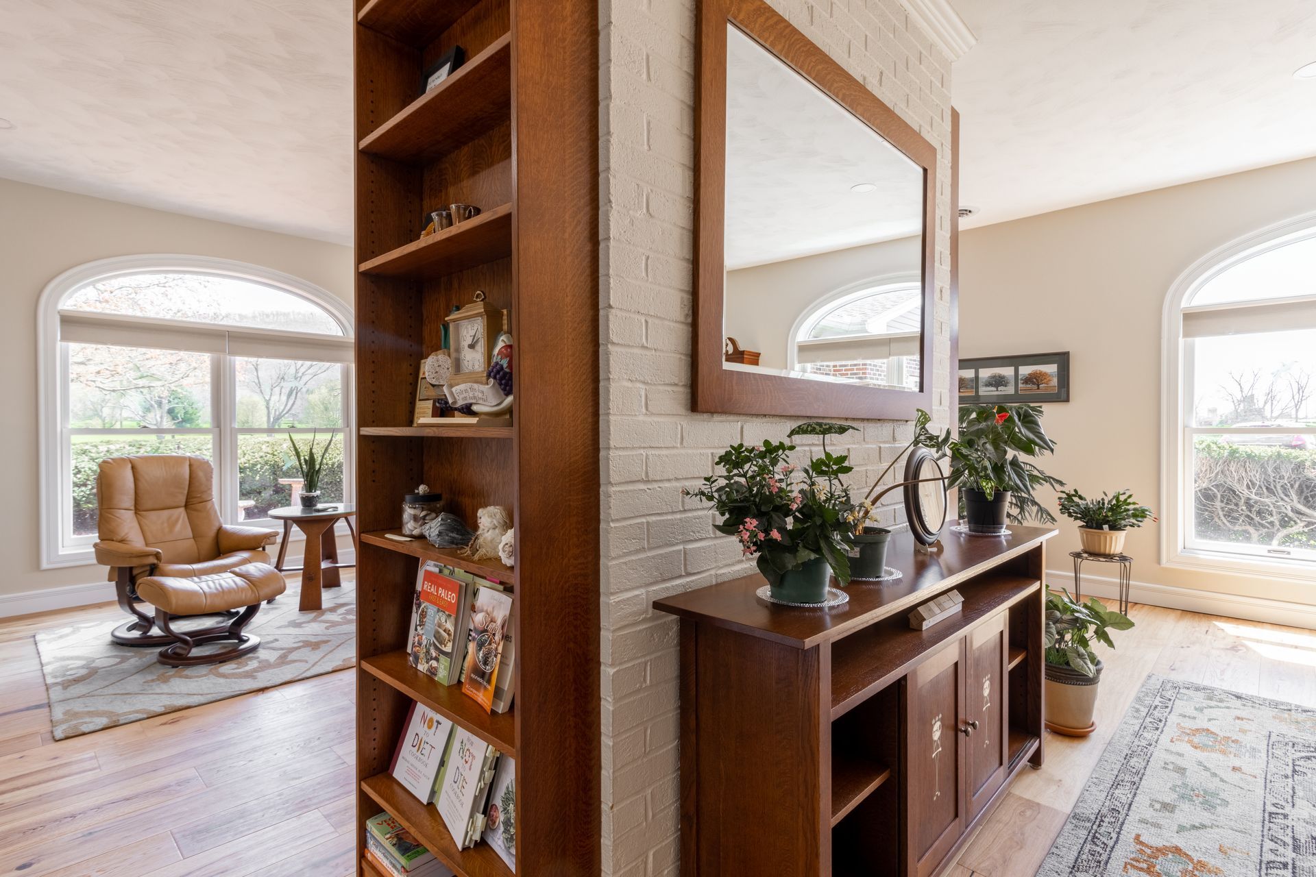 Interior with bookshelf, recliner, console table, arched windows, and hardwood floors.