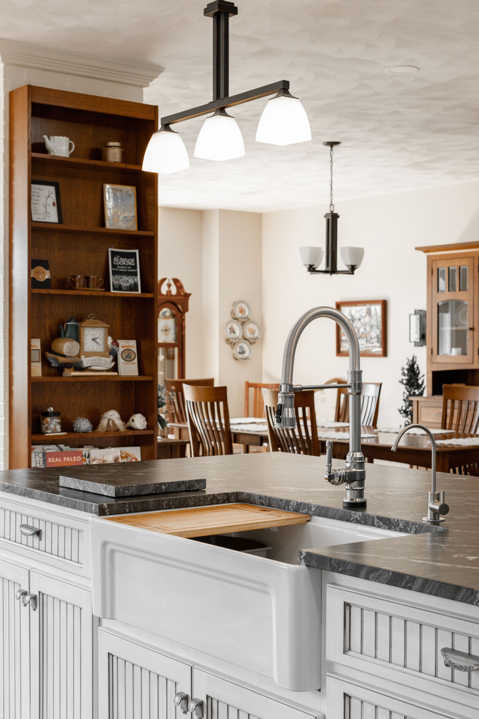 Kitchen with farmhouse sink, countertop, and pendant lights. Wooden hutch and dining table visible in the background.
