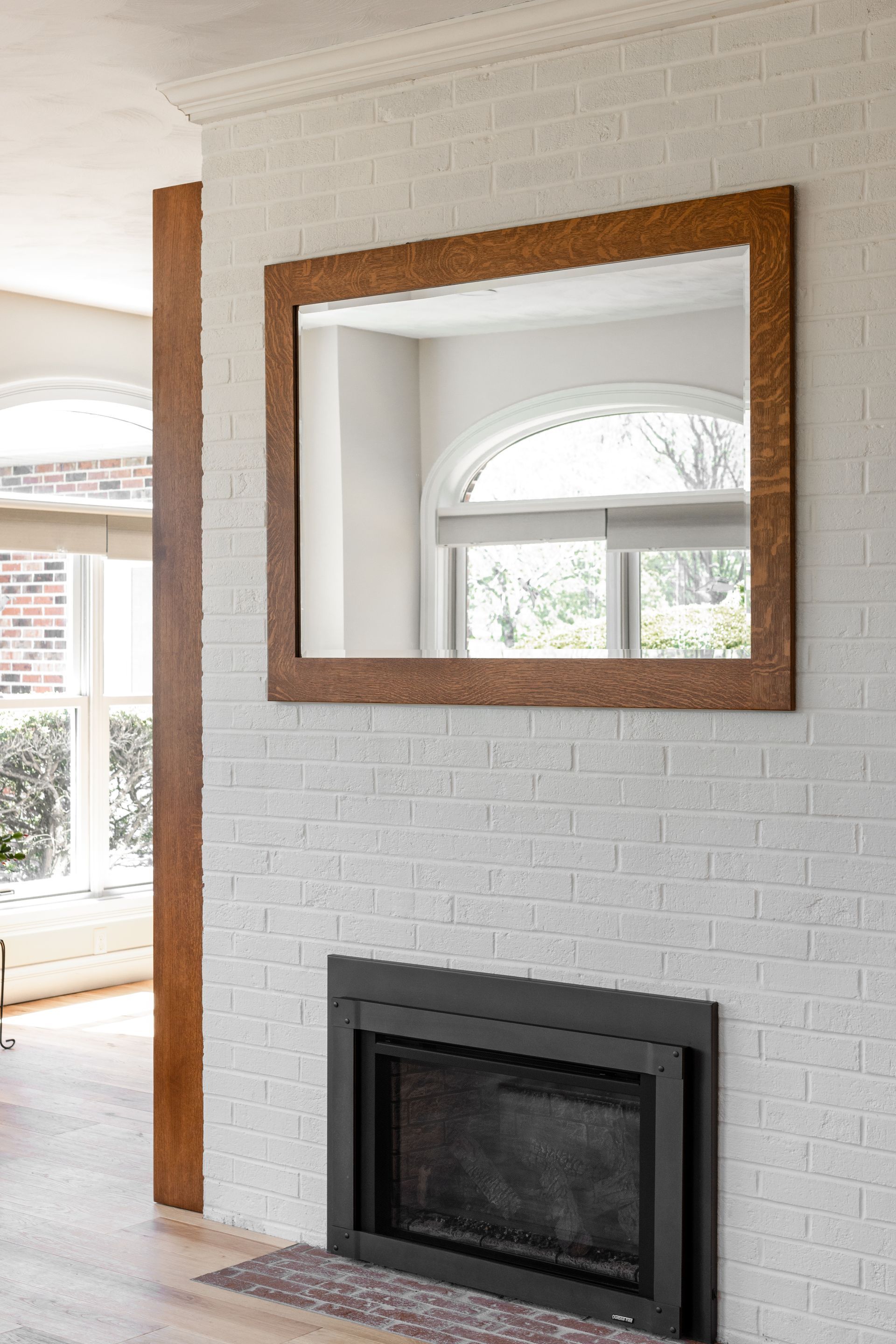 Mirror above black fireplace on white brick wall. Brown trim and a large window is reflected.