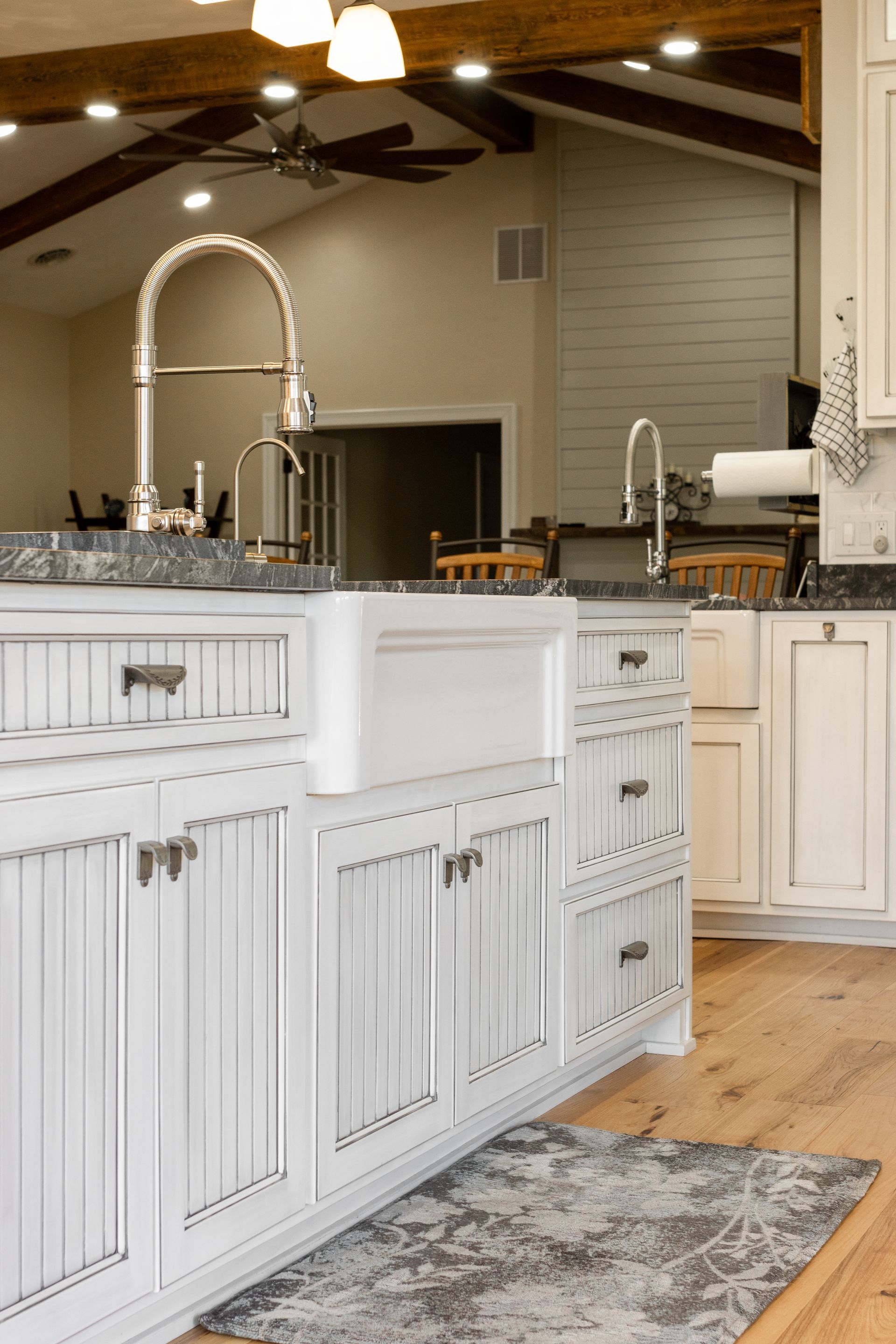 White kitchen island with a farmhouse sink, light wood floor, and exposed beam ceiling.