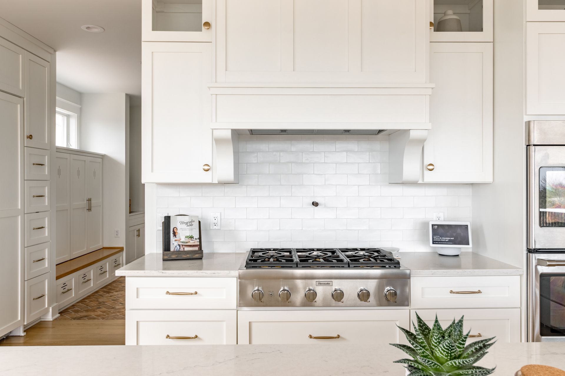 White kitchen with range, cabinets, and light countertop. Subtile tile backsplash, and small plant.