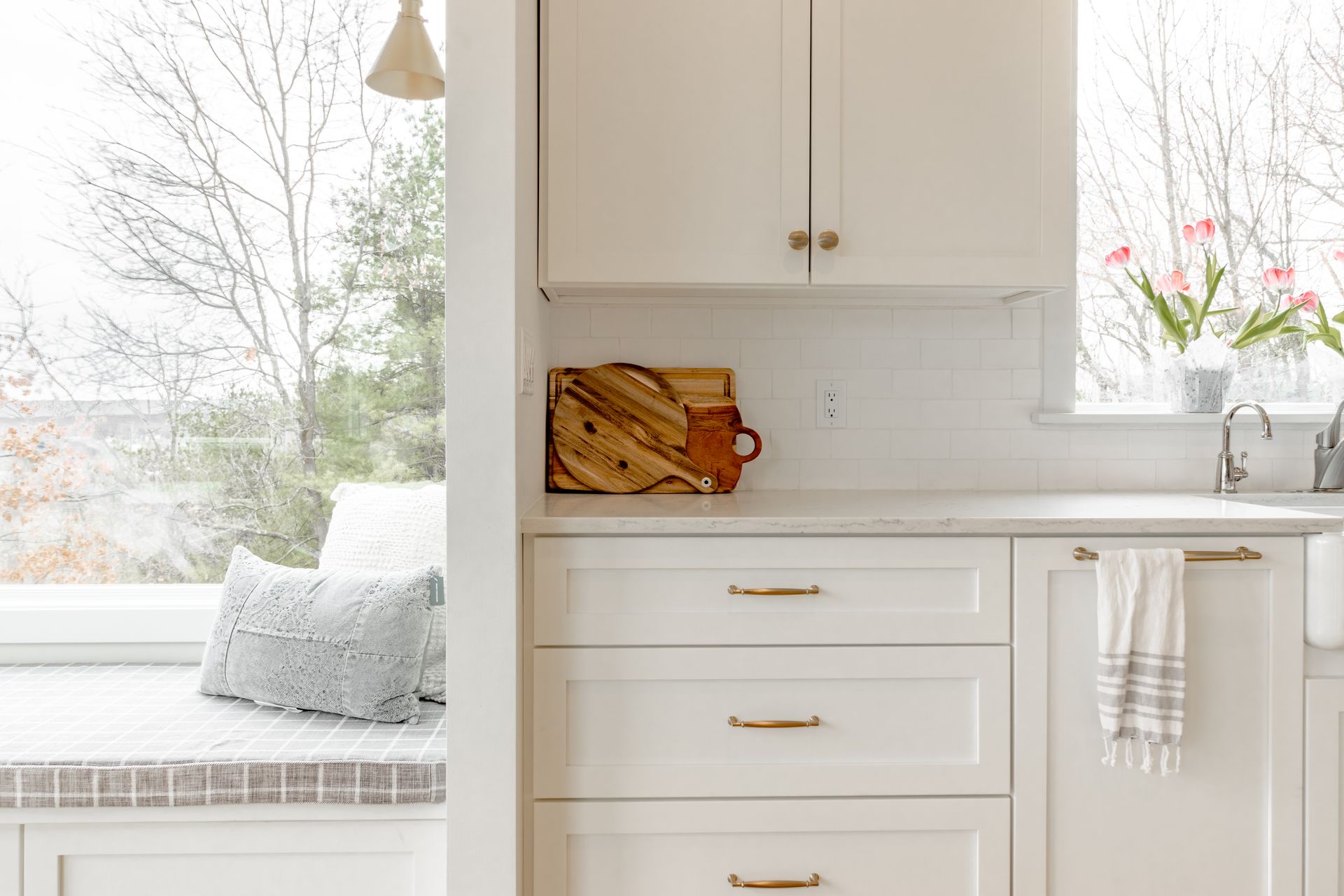 White kitchen cabinets with gold hardware, wooden cutting board, sink, and window seat with a pillow.