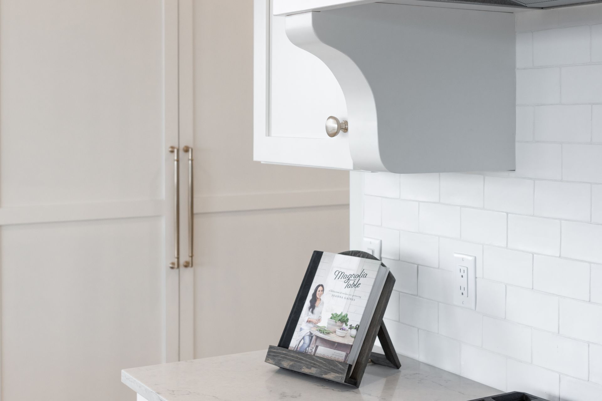 White kitchen with cookbook on counter. Gray cabinet, white tile backsplash, and light gray countertop.