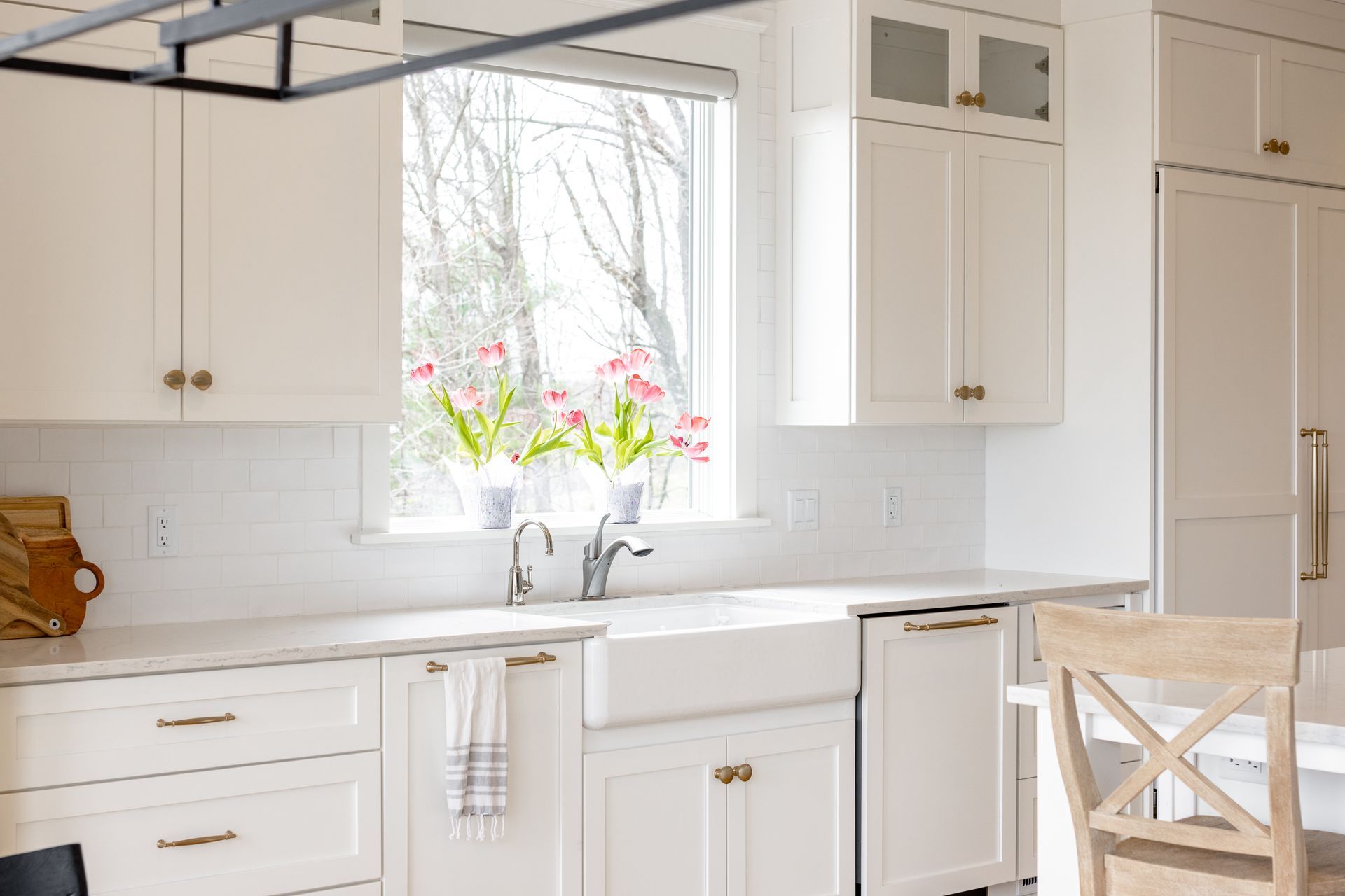 White kitchen with a farmhouse sink, gold hardware, and pink tulips in the window.