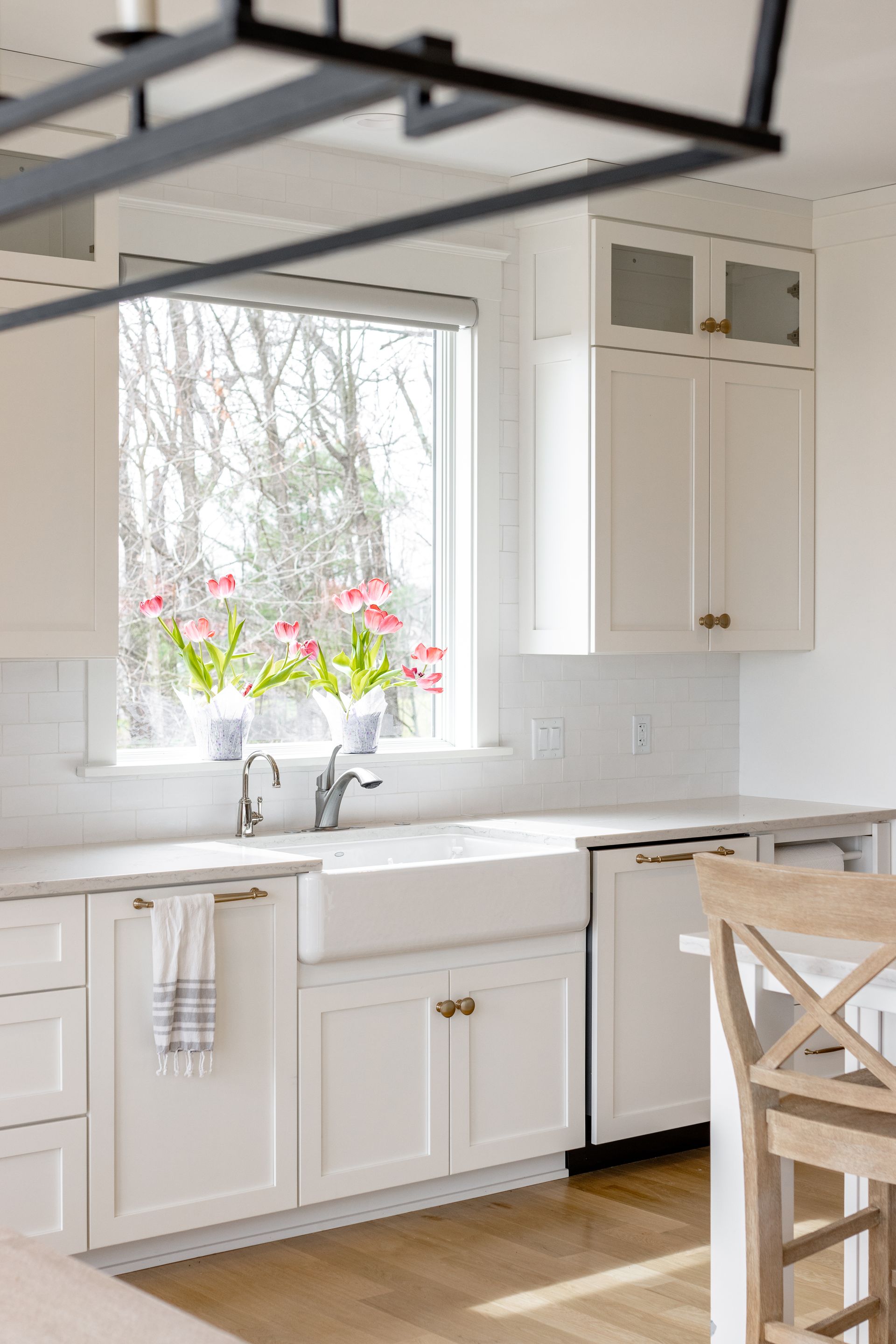 White kitchen with window over farmhouse sink, flowers, cabinets, and light wood floor.