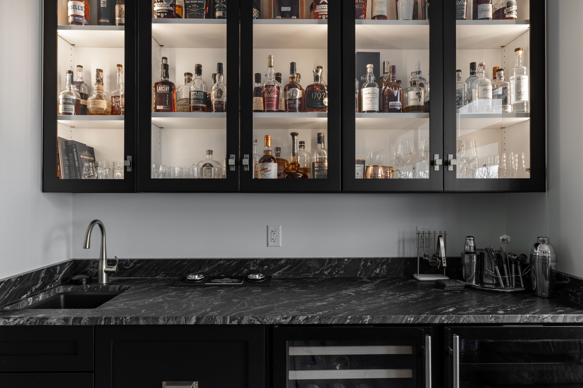 A built-in bar with black cabinets, glass doors showcasing liquor bottles, and a granite countertop.