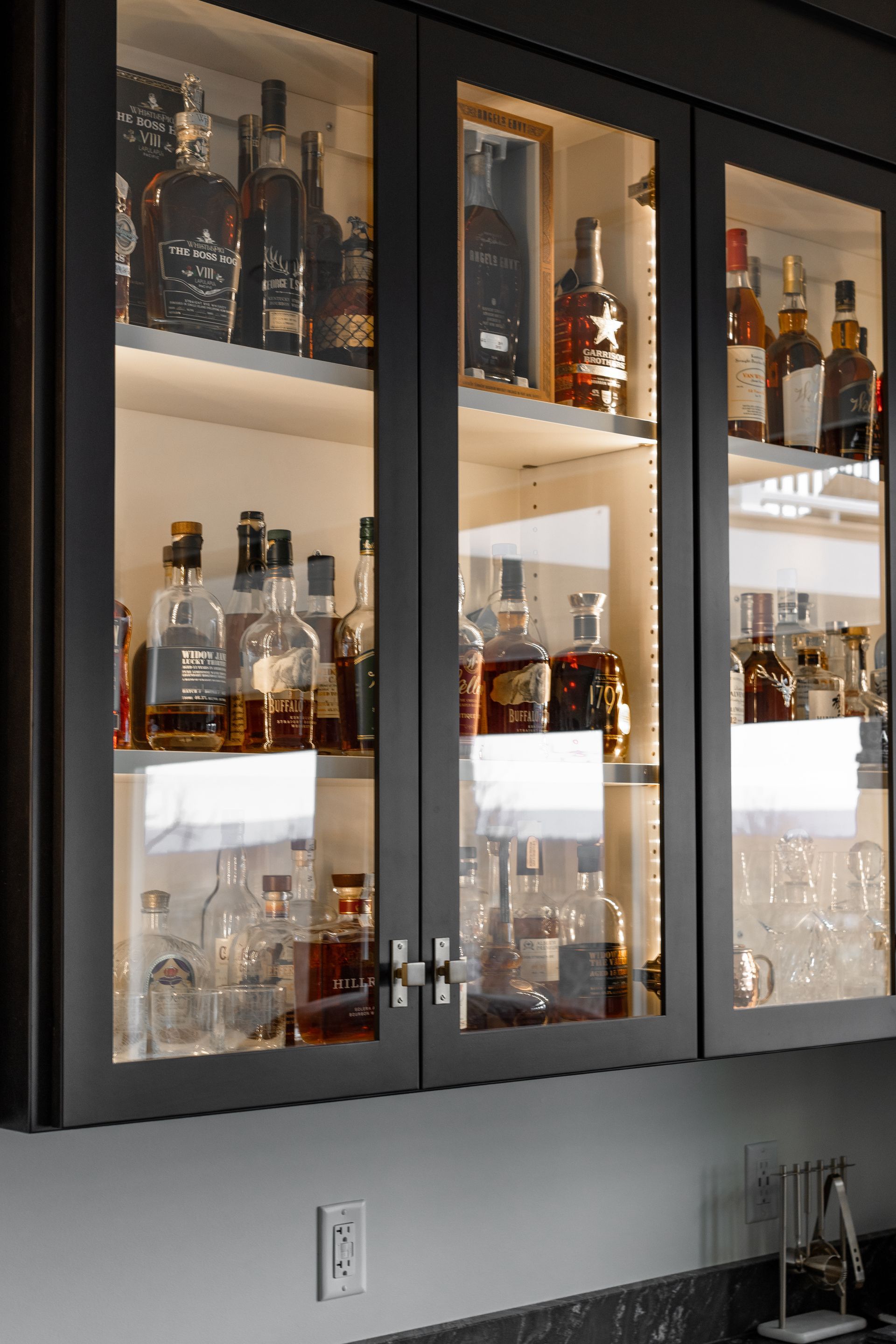 Lit glass-front liquor cabinet with bottles. Dark gray cabinets against a white wall.