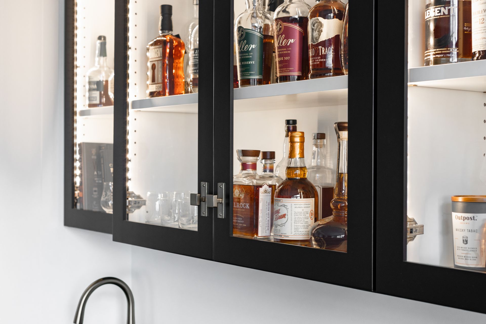 Black liquor cabinets with glass doors, illuminated interior, stocked with bottles. Mounted above a sink.