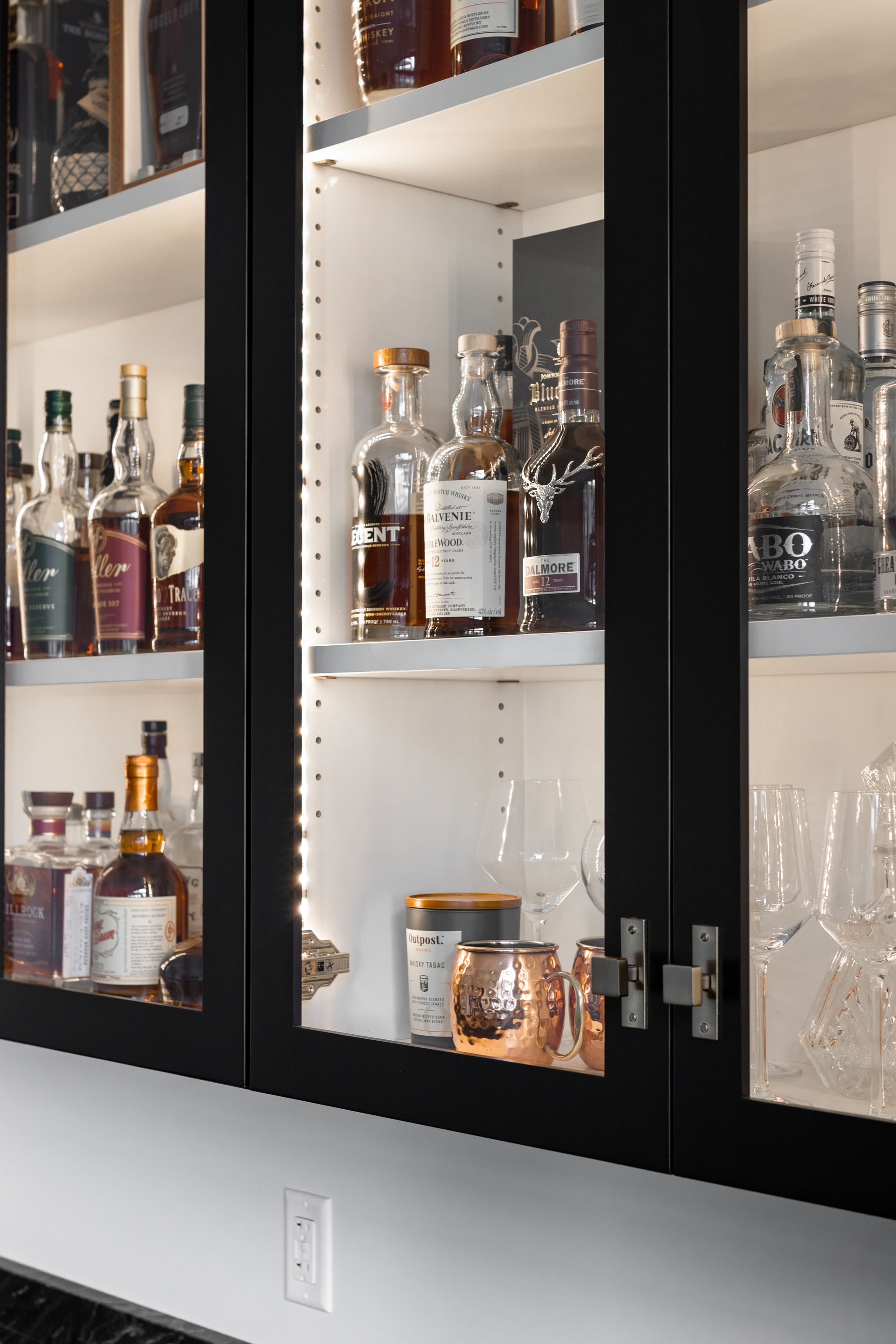 Black cabinet with liquor bottles, glasses, and copper mugs on lit shelves.