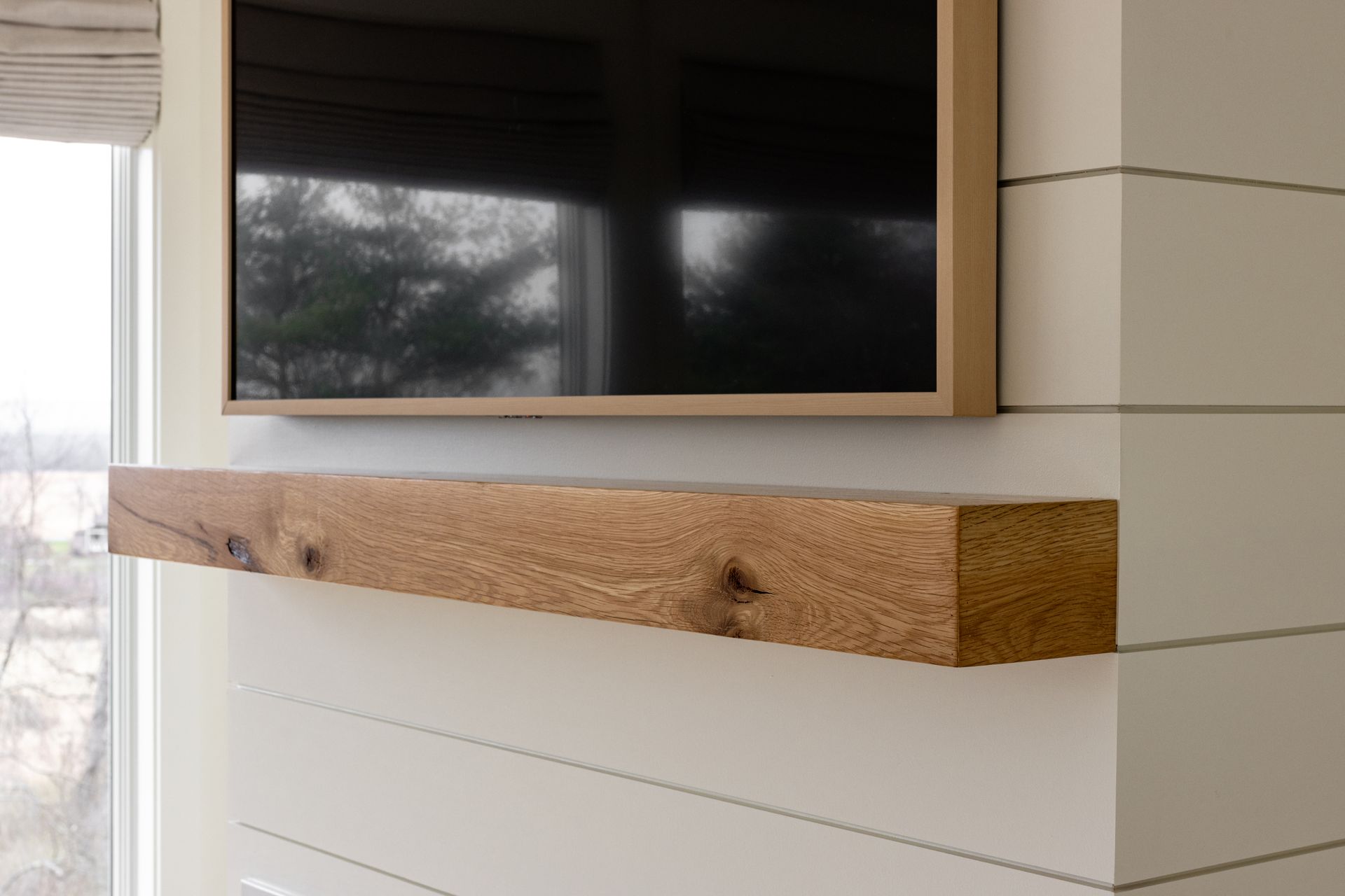 Wooden shelf below a mounted television on a white paneled wall with a window.