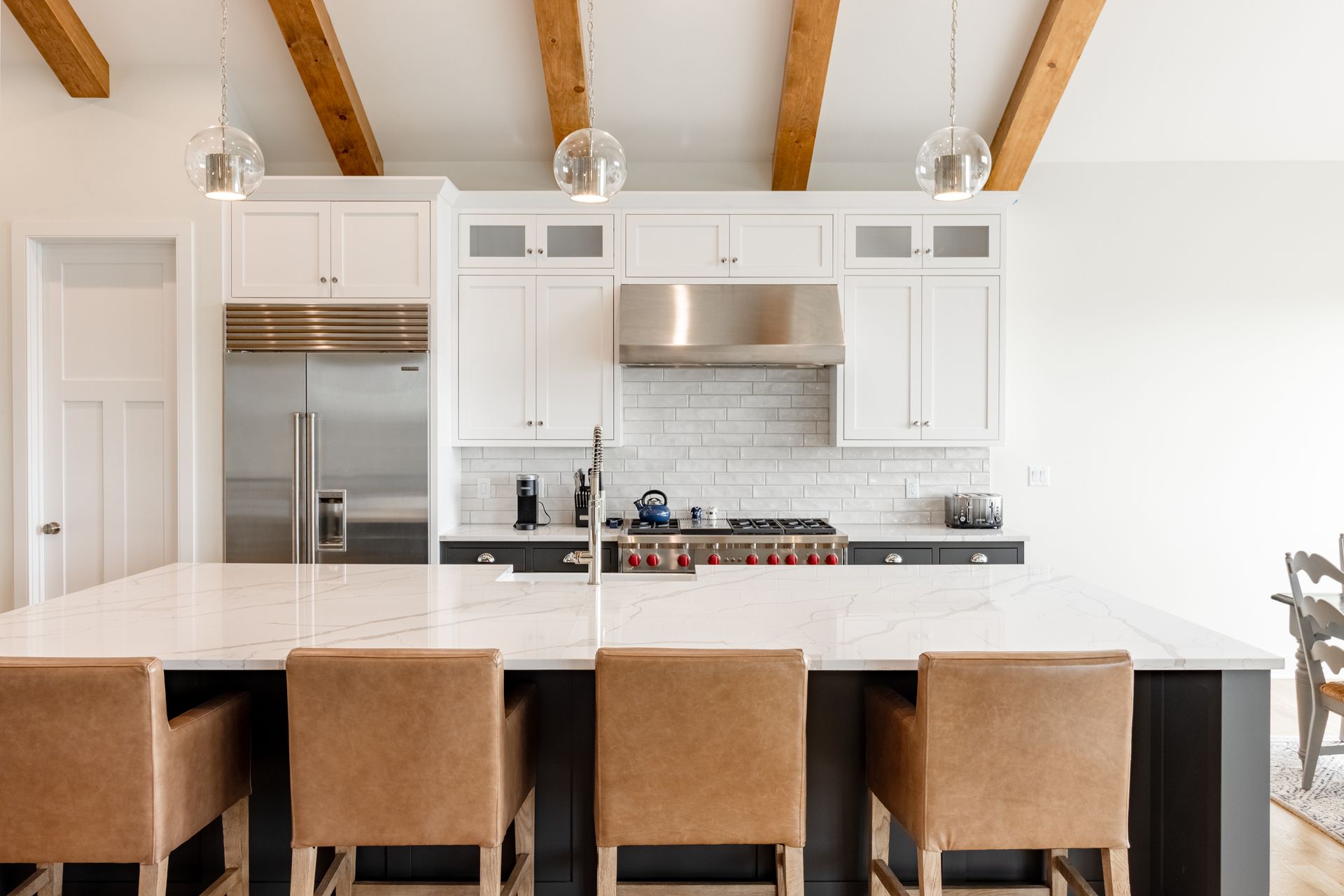 Modern kitchen with white cabinets, large island, brown leather stools, and wood beams.