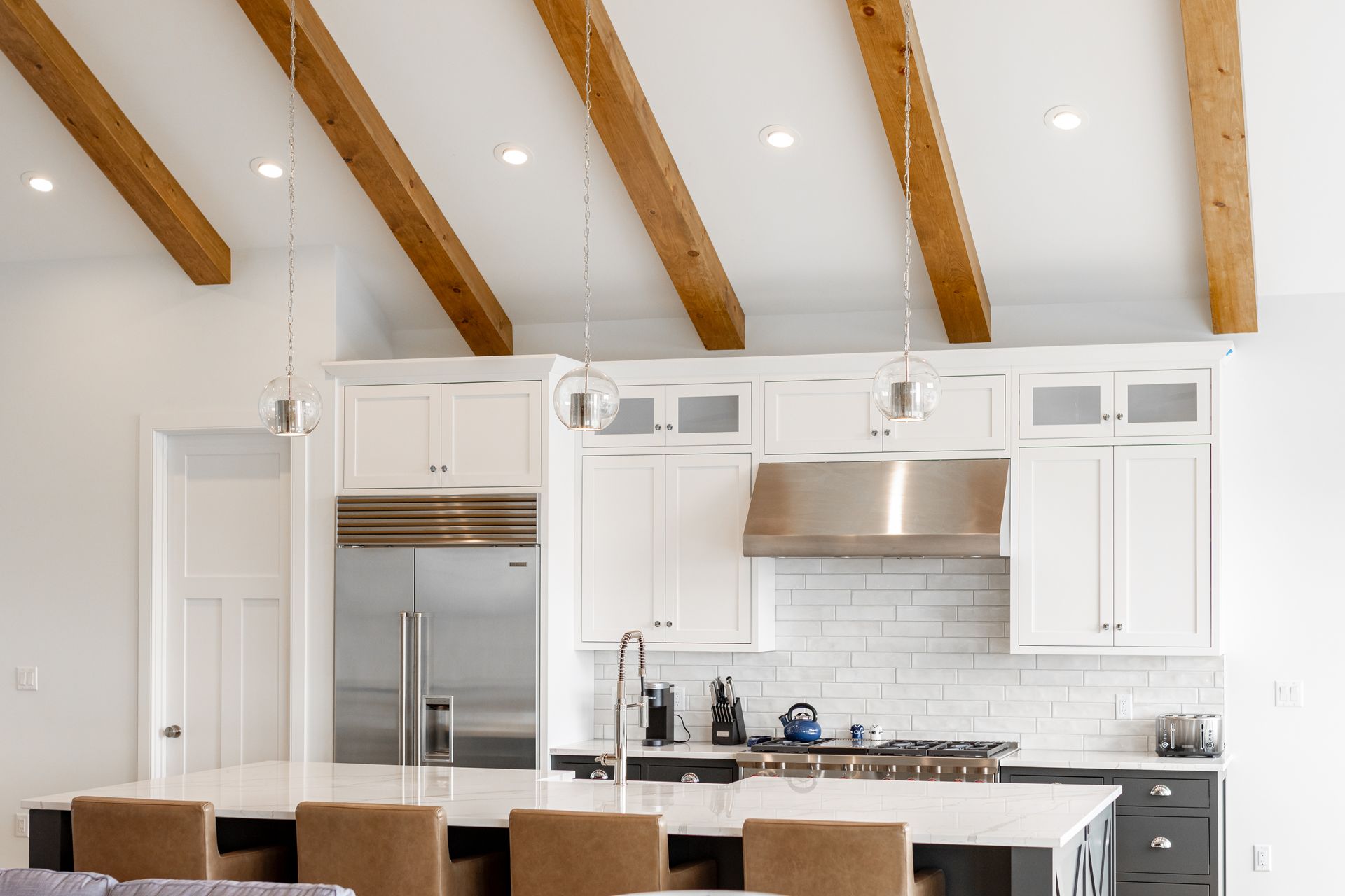 White kitchen with island, stainless appliances, exposed wooden beams, and pendant lights.