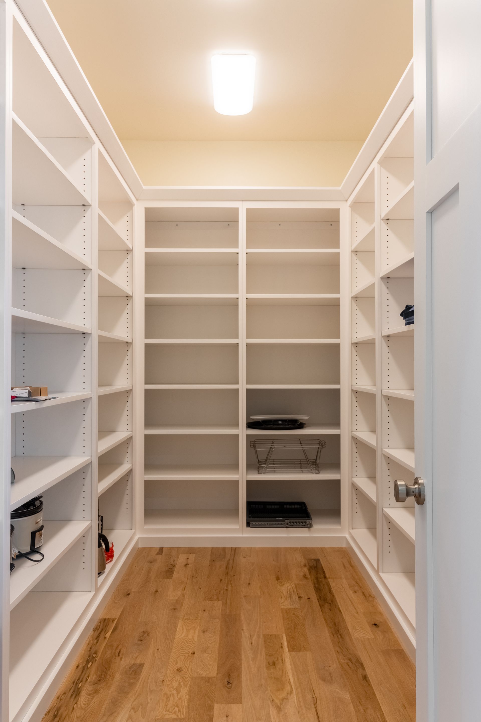 Empty white pantry with multiple shelves, wood flooring, and a light fixture.