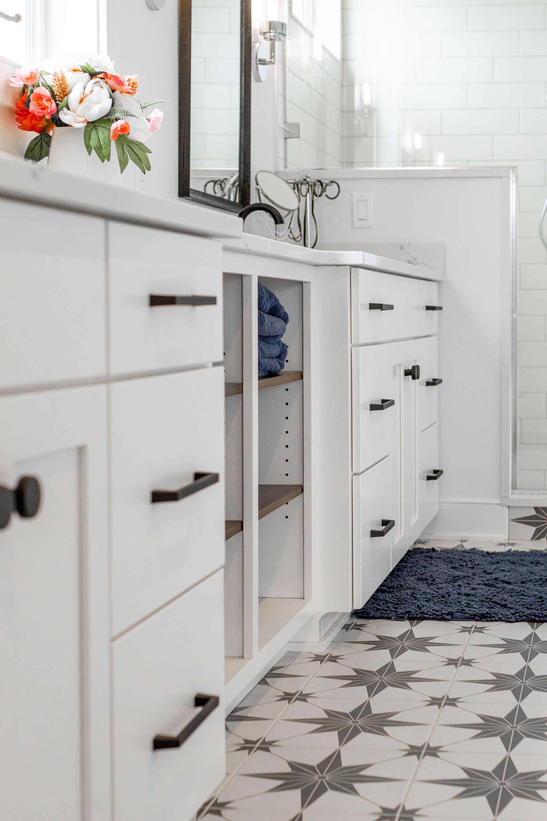 White bathroom vanity with black hardware. Open cabinet with blue towels, star-patterned tile floor.