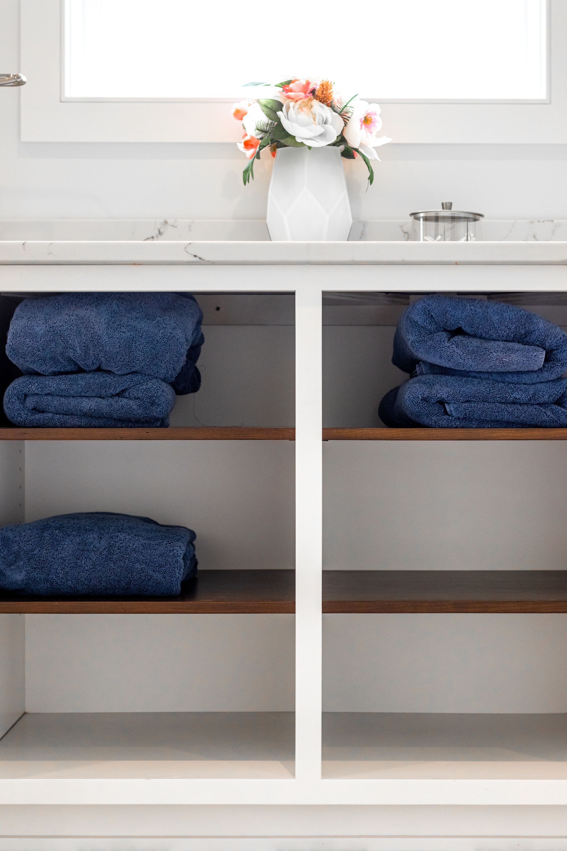 White cabinet with stacked blue towels and a vase of flowers on a countertop.