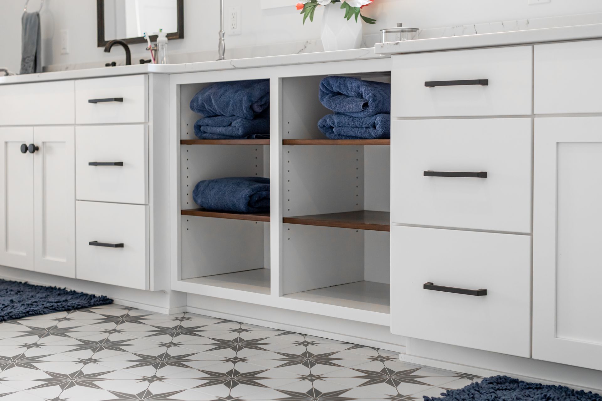 White bathroom vanity with open shelving holding blue towels; patterned tile floor.
