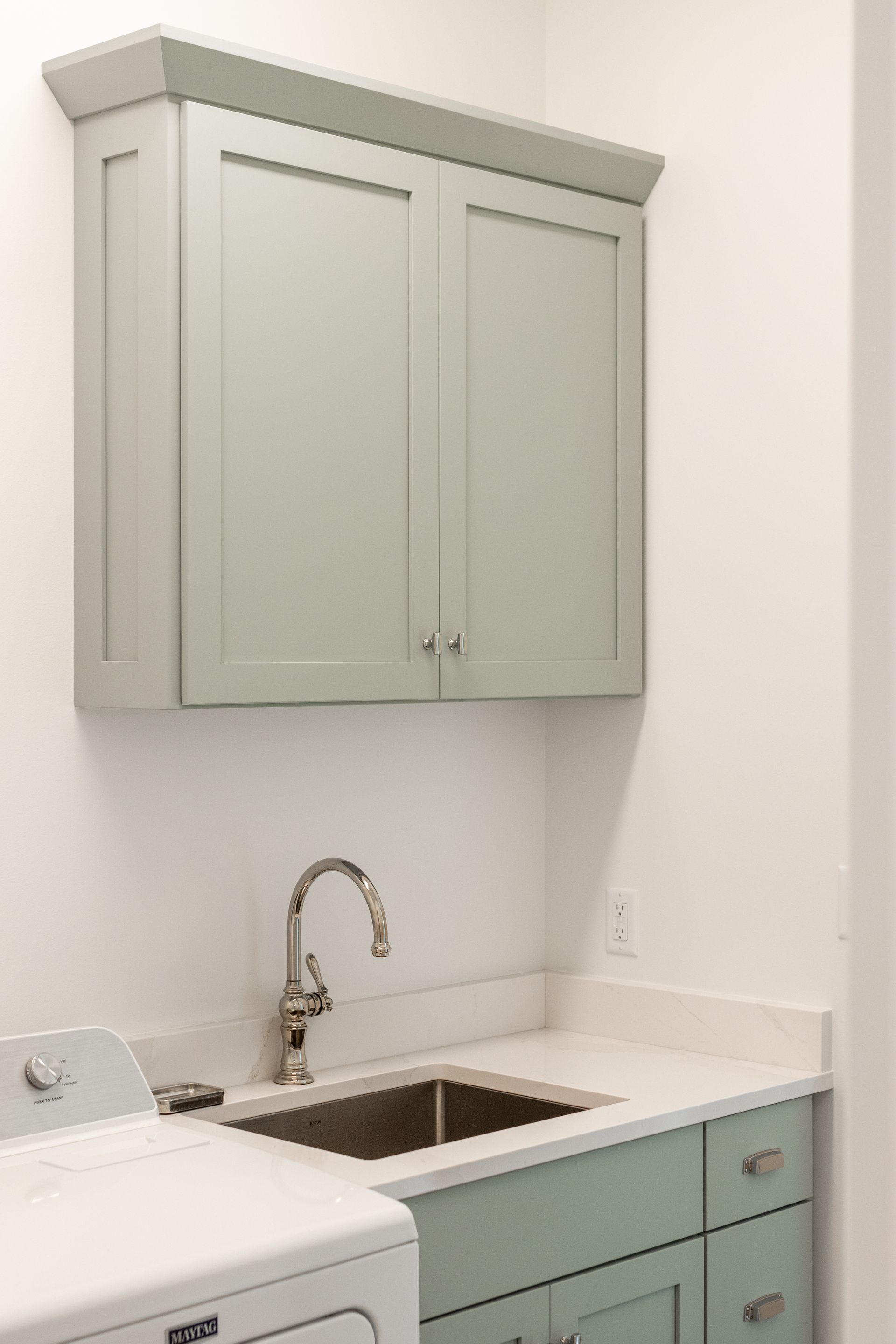 Light green cabinetry over a sink in a laundry room, with a faucet, and a washing machine.