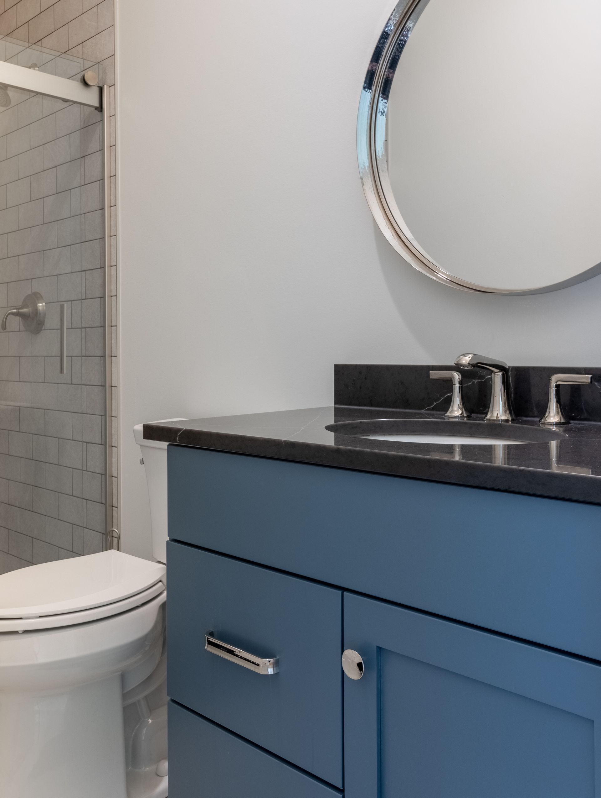 Blue bathroom vanity with dark countertop, round mirror, and toilet.
