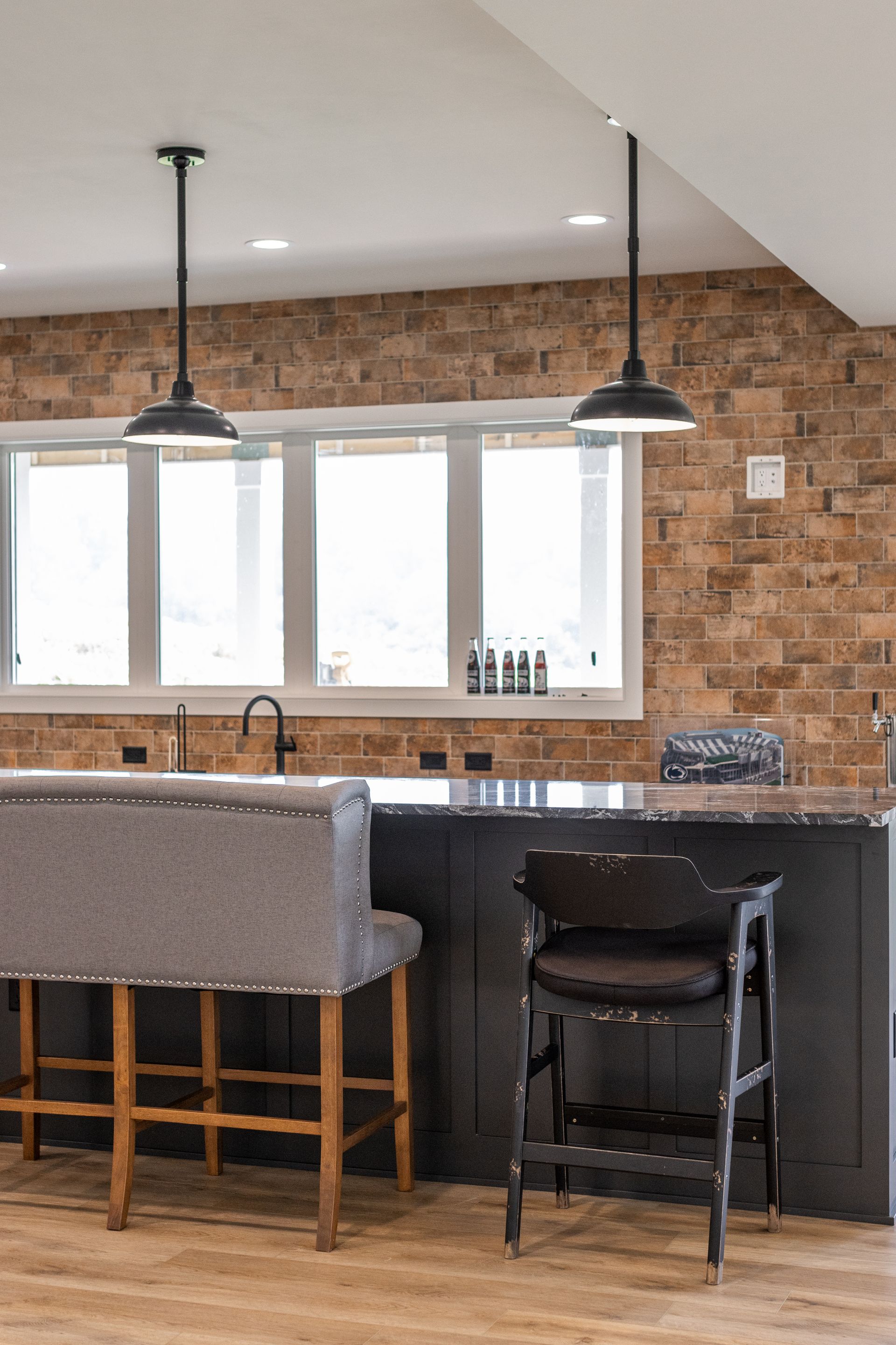 Kitchen island with a gray upholstered bench, dark barstool, brick wall, and two pendant lights.