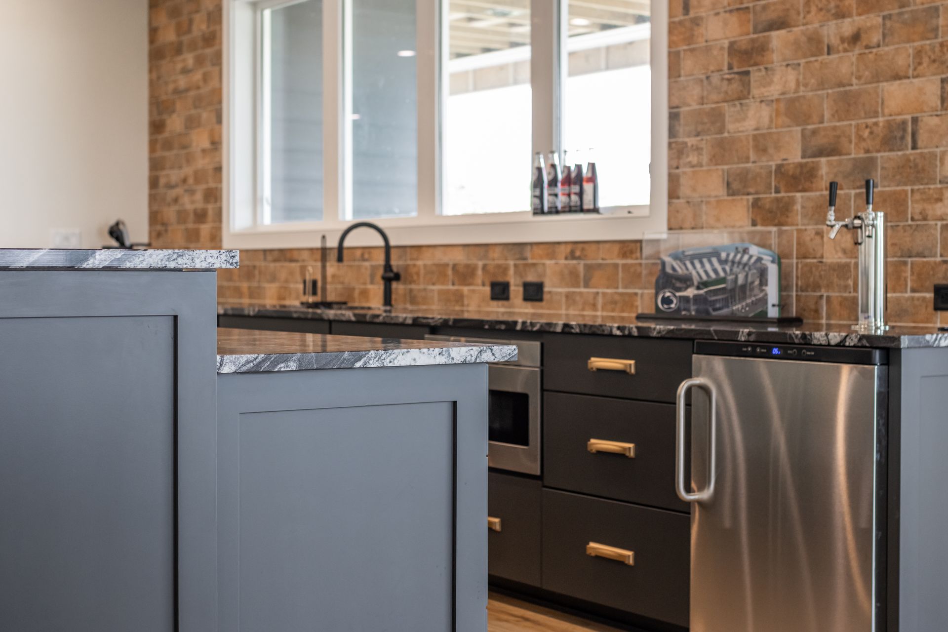 Bar area with gray cabinets, a small microwave, and a stainless steel fridge. Brick tile backsplash and window.