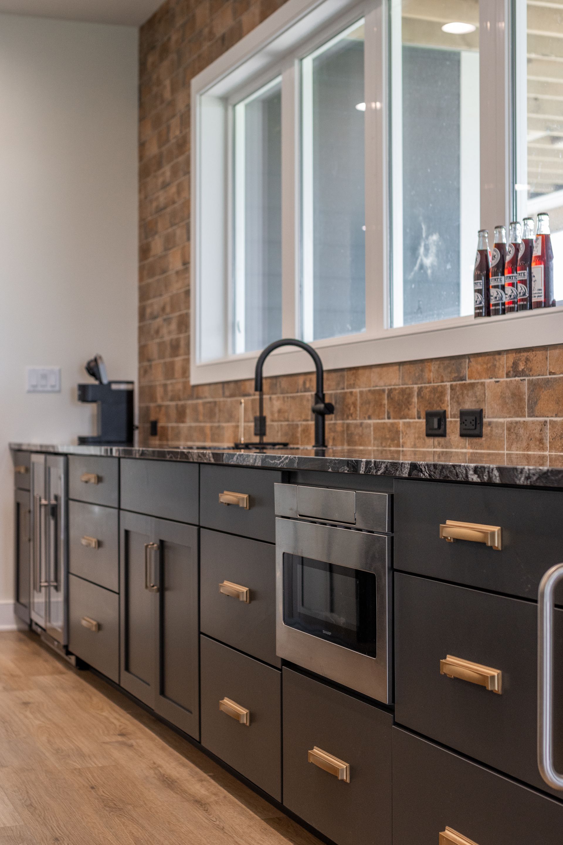 Dark cabinets with gold hardware in a kitchen, black countertop, brick backsplash, window, and a stainless steel microwave.