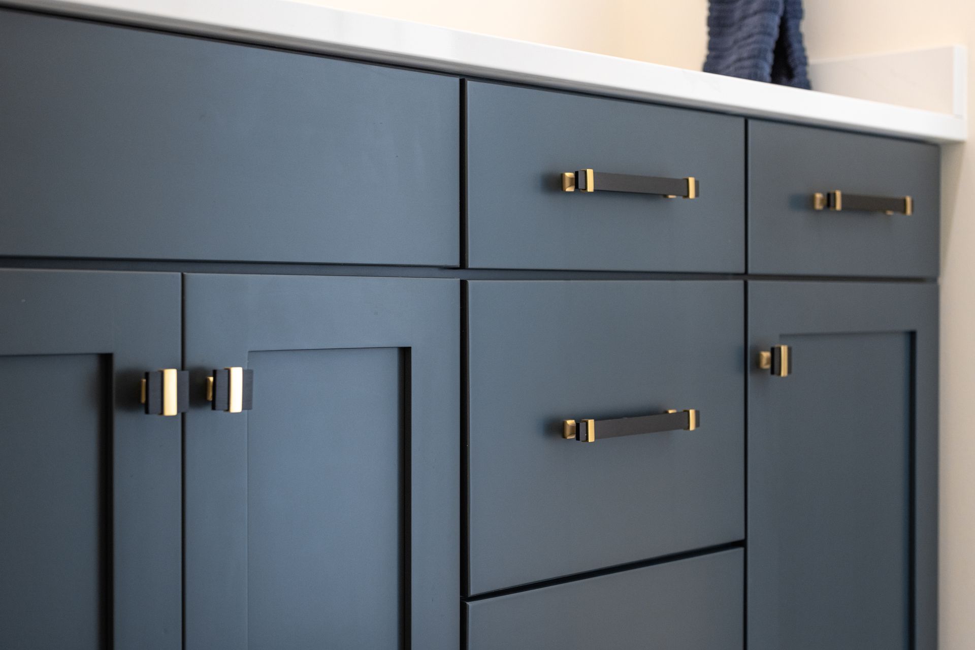 Blue bathroom cabinets with gold and black hardware, below a white countertop.