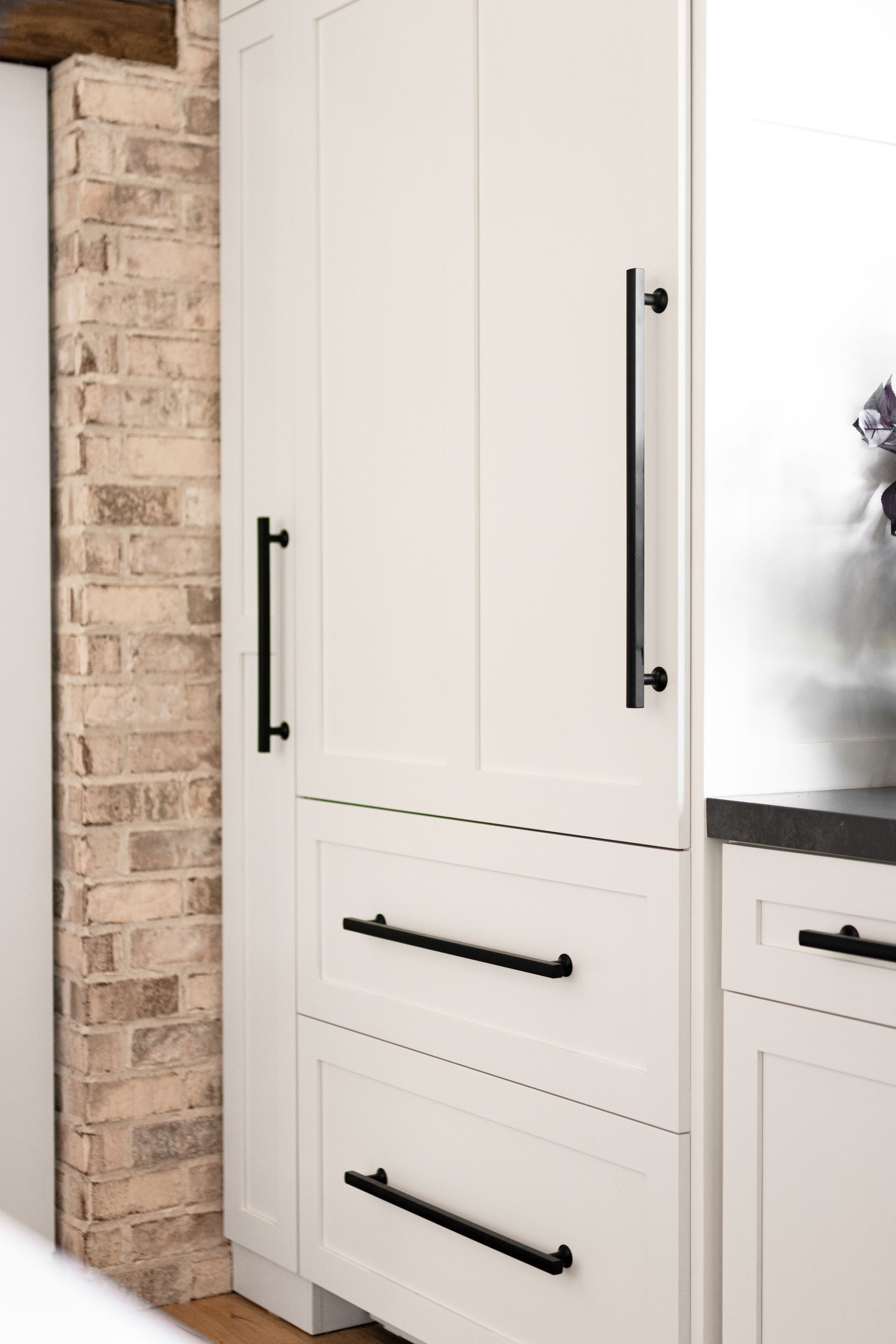 White kitchen cabinets with black handles against a brick wall.