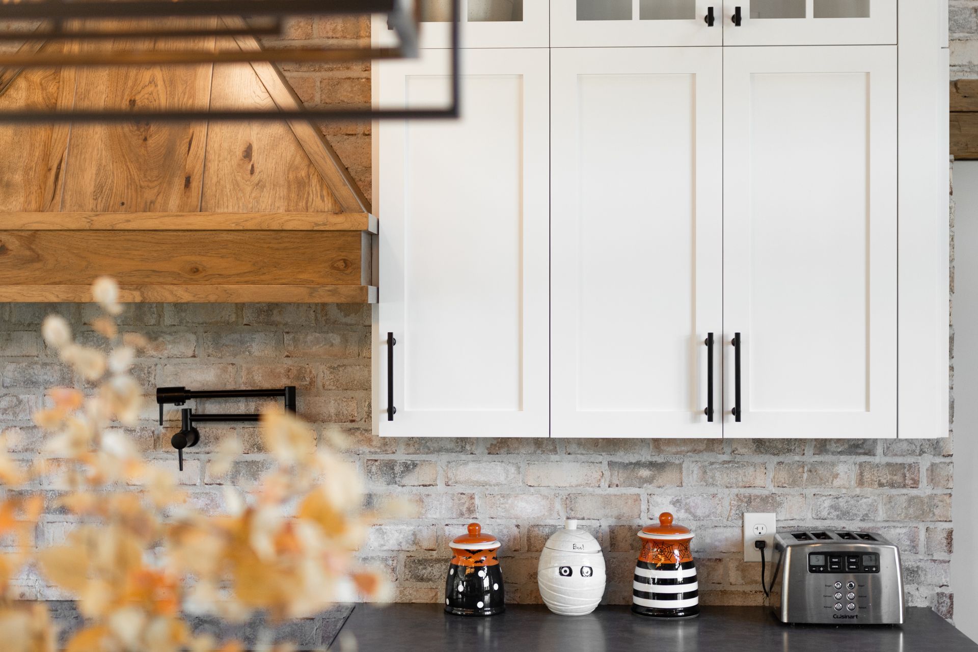 White kitchen cabinets with black handles, brick backsplash, and a wooden range hood.