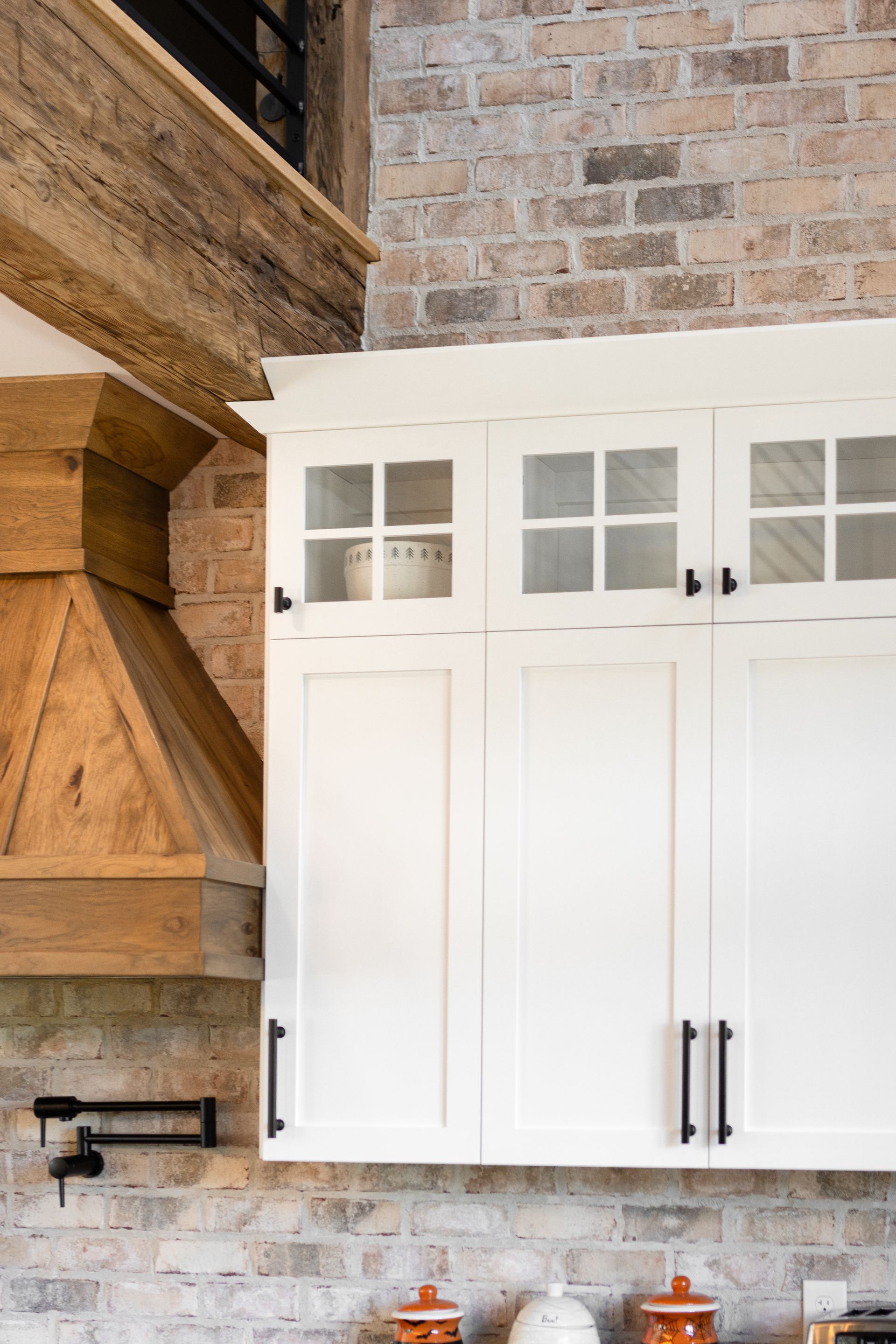 White kitchen cabinets with glass-paned uppers against a brick wall. A wood hood and beam are visible.