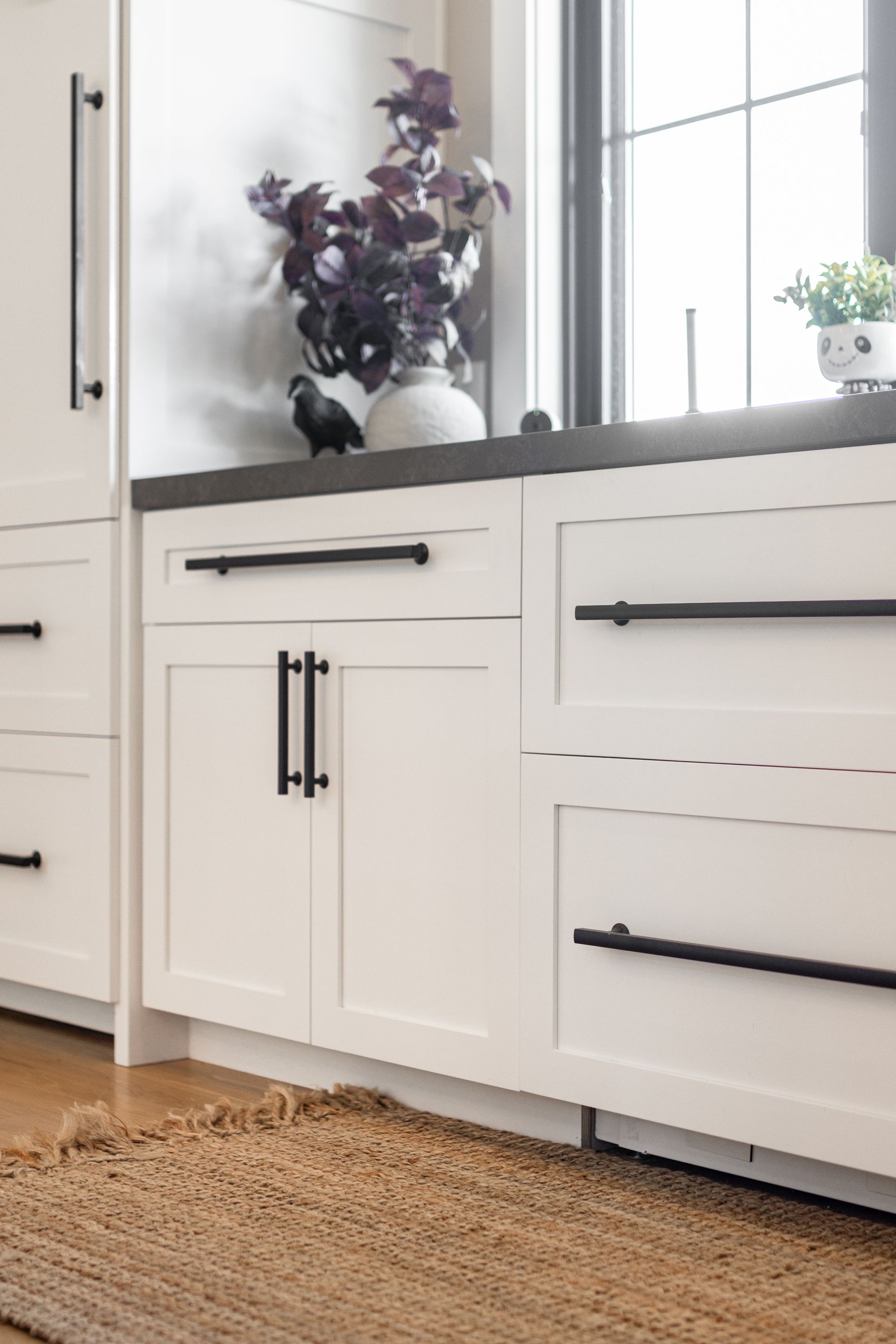 White kitchen cabinets with black hardware, next to a window, rug in foreground.