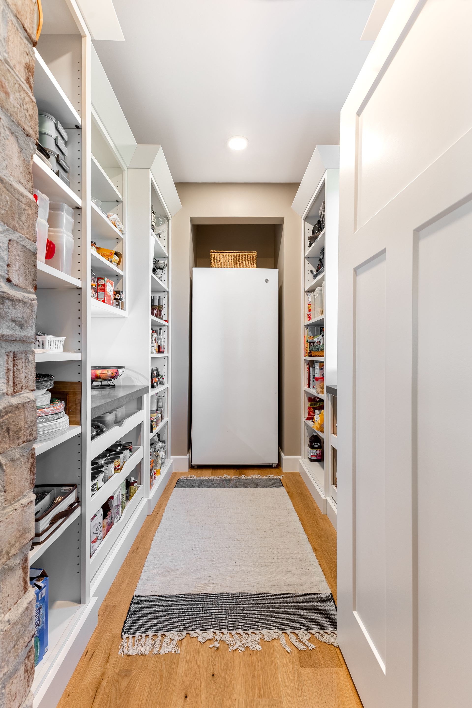 Narrow pantry with shelves, brick wall, refrigerator, and rug. Wooden floor, white cabinetry.