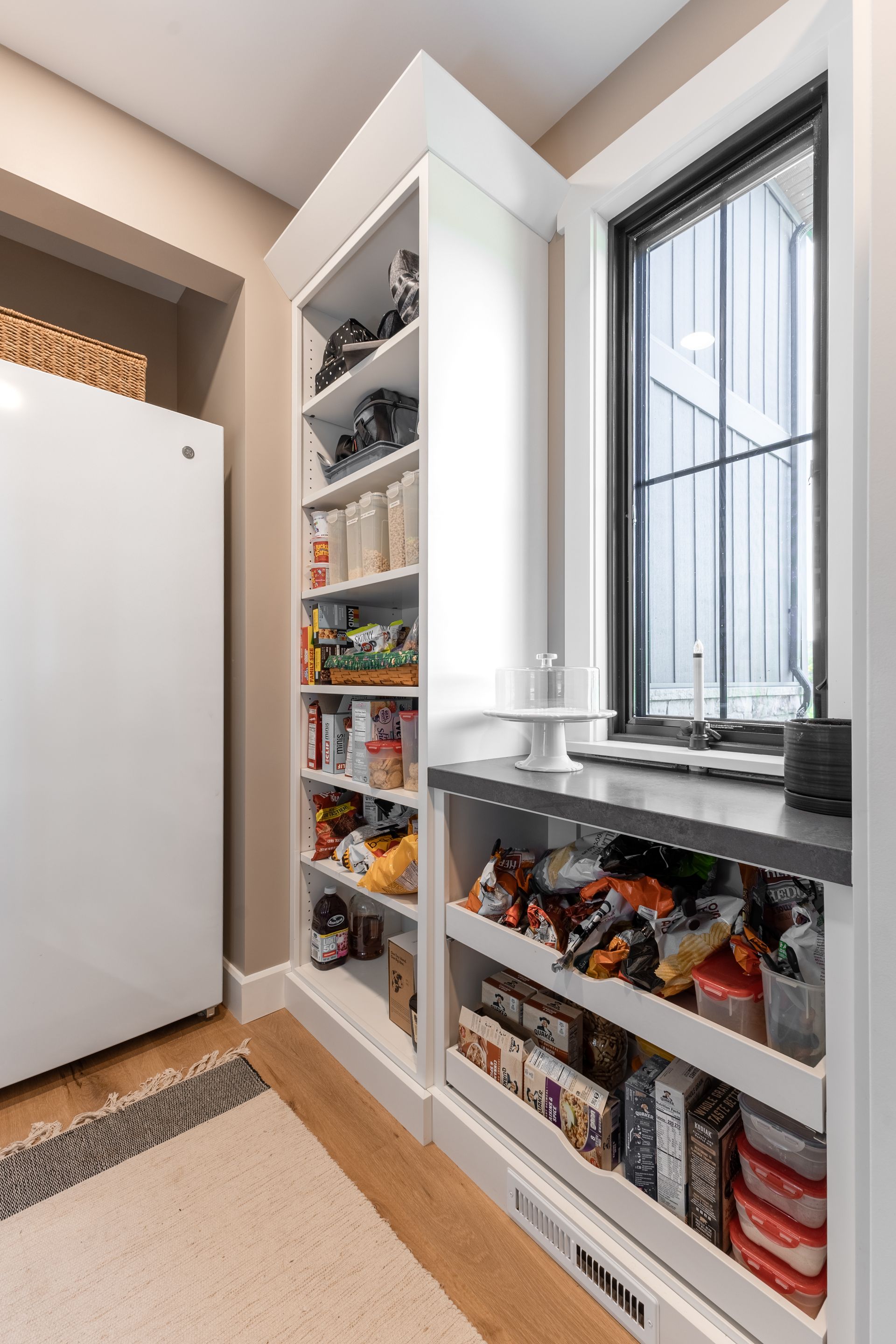 White pantry with open shelves, adjacent to a window. Snacks and food items are visible.