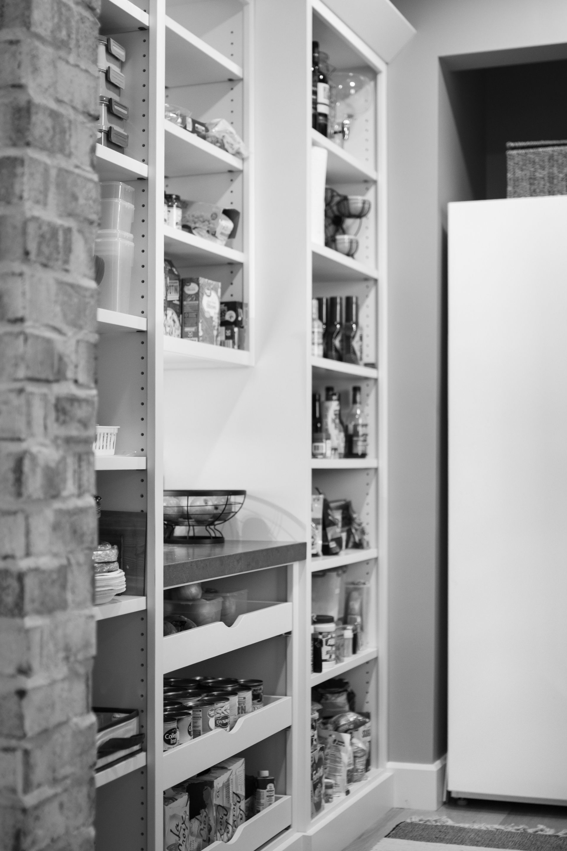 A narrow pantry with white shelves filled with food items next to a brick wall and a refrigerator.