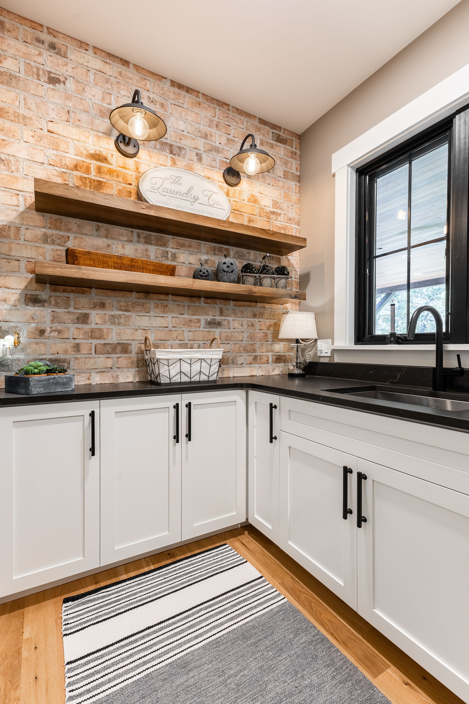 Kitchen with white cabinets, brick wall, and black countertops; wooden shelves with decorations and two sconces.