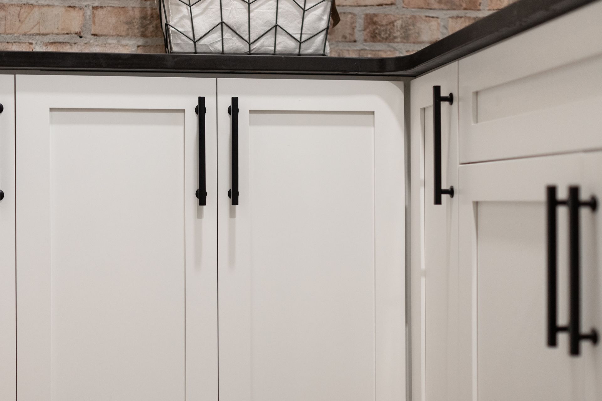 White kitchen cabinets with black handles against a brick wall.