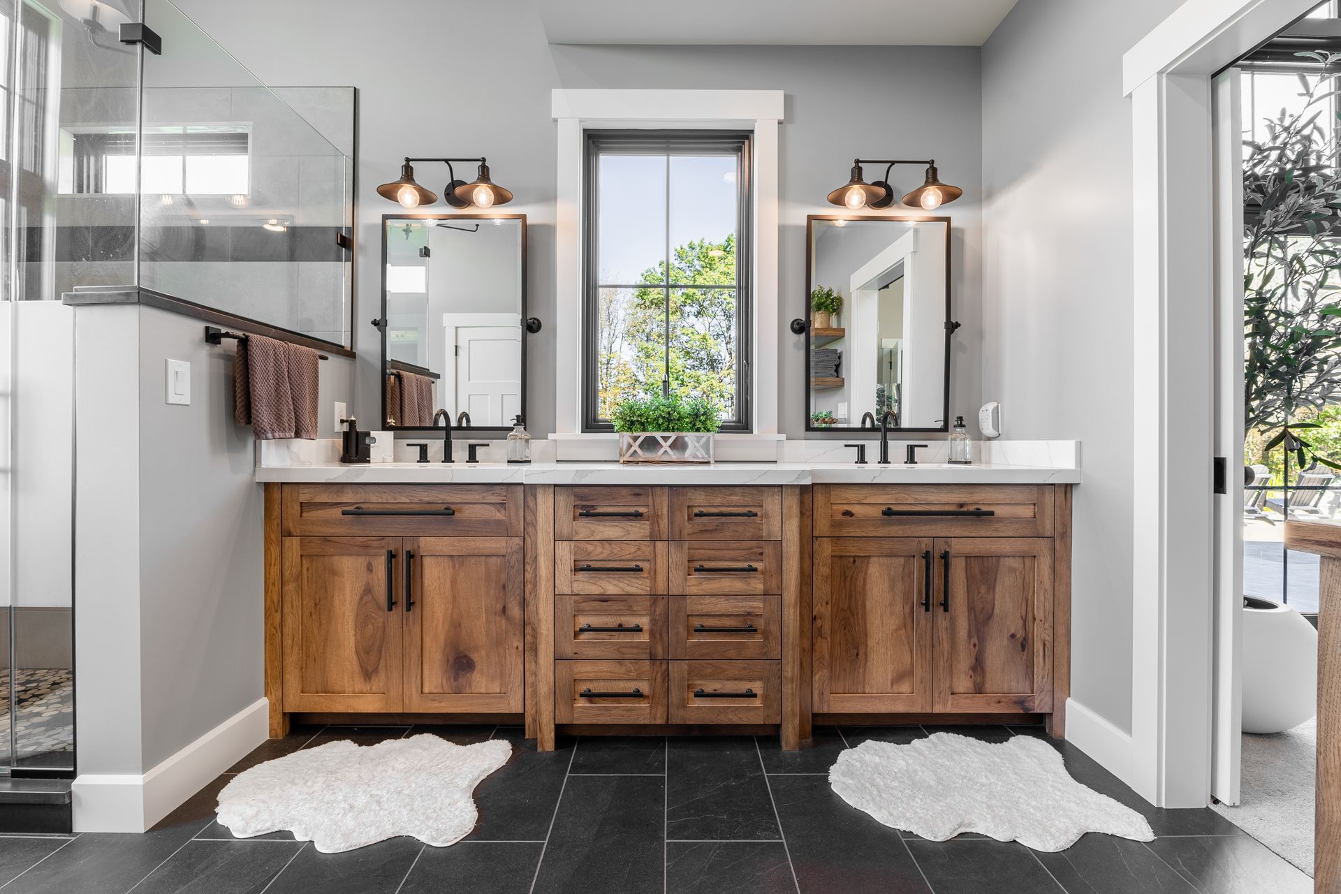 Bathroom with wood vanity, black countertops, mirrors, and a window; two white rugs on dark tiled floor.
