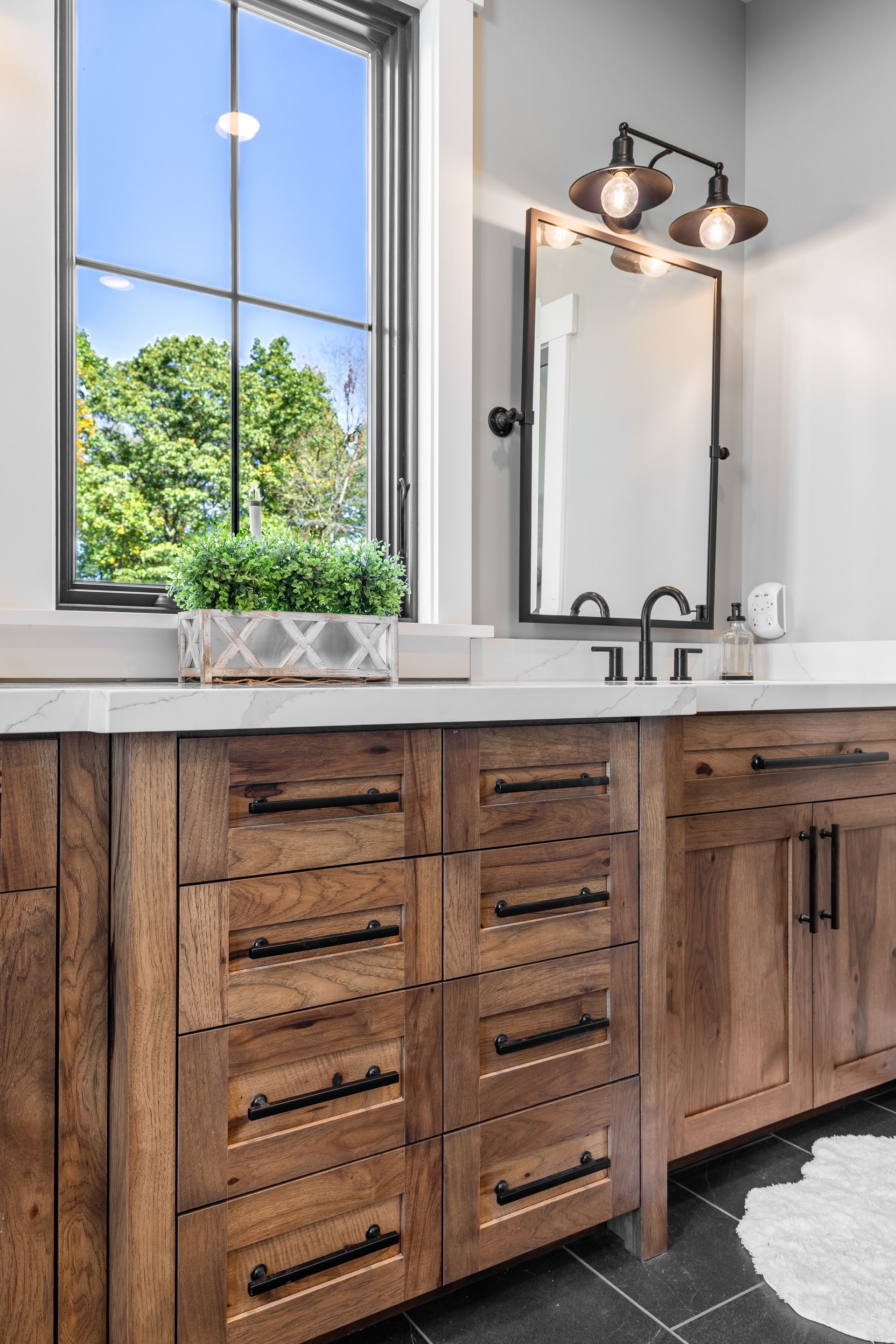 Bathroom vanity with wood cabinets, black hardware, window, mirror, and light fixture.