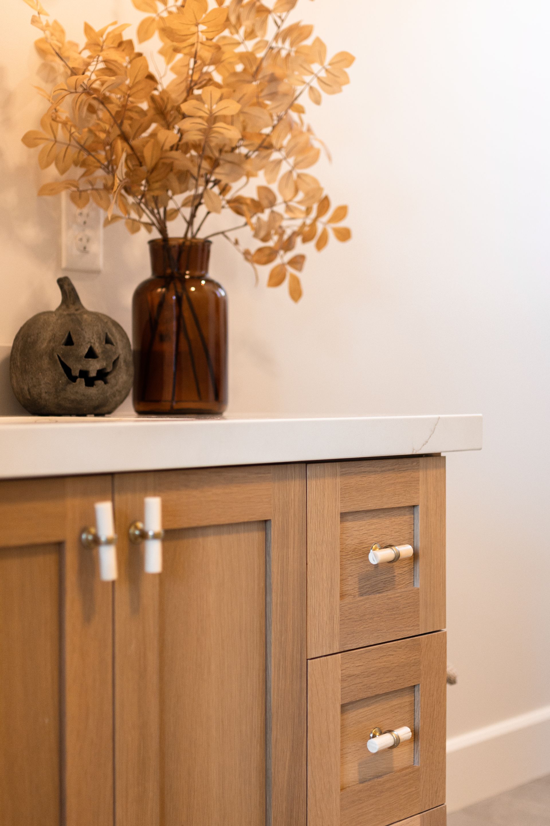 Wooden cabinet with white countertop, brown vase with yellow branches, a carved pumpkin.