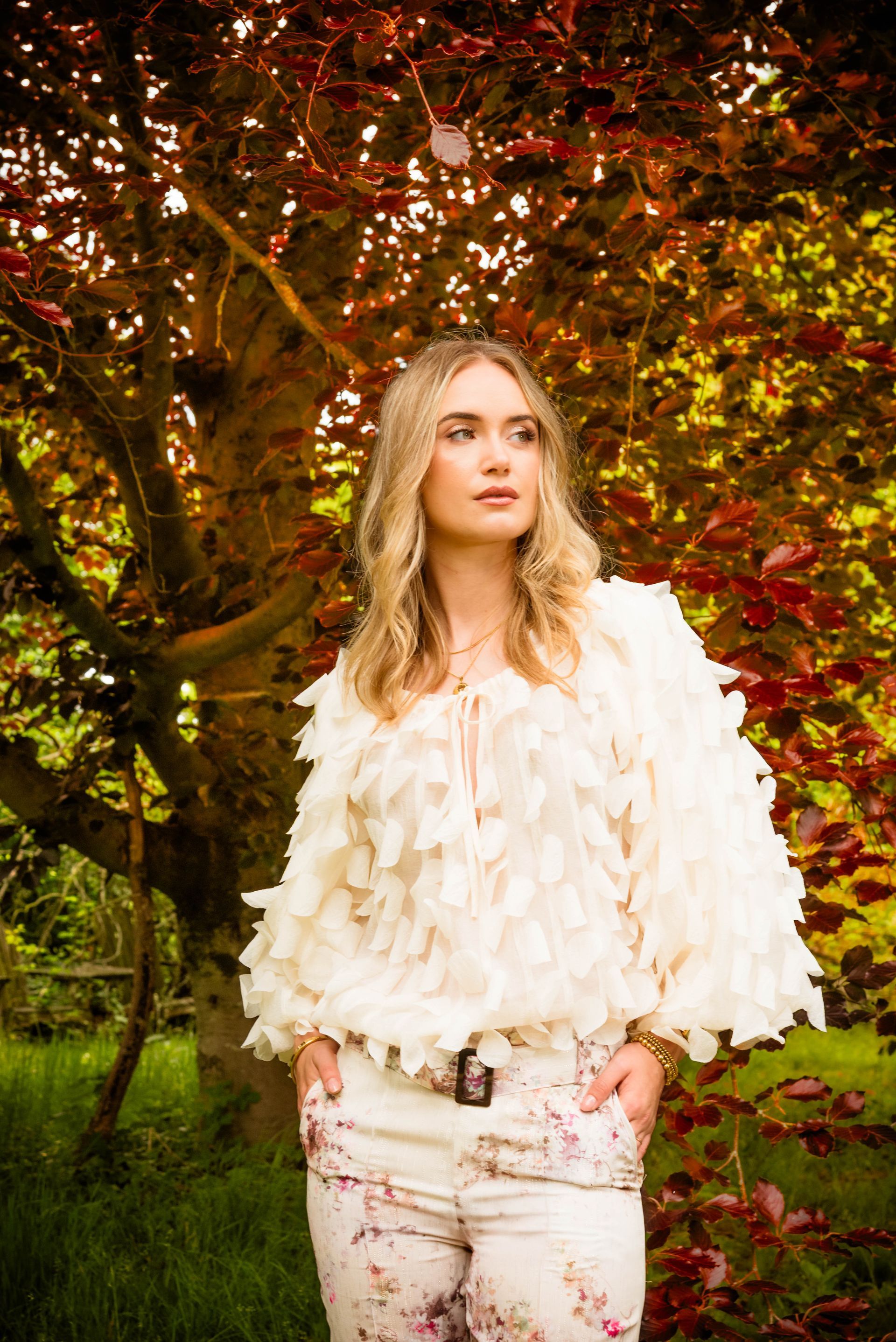 Vrouw in crèmekleurige top en bloemenbroek, staand voor een boom met rode bladeren.