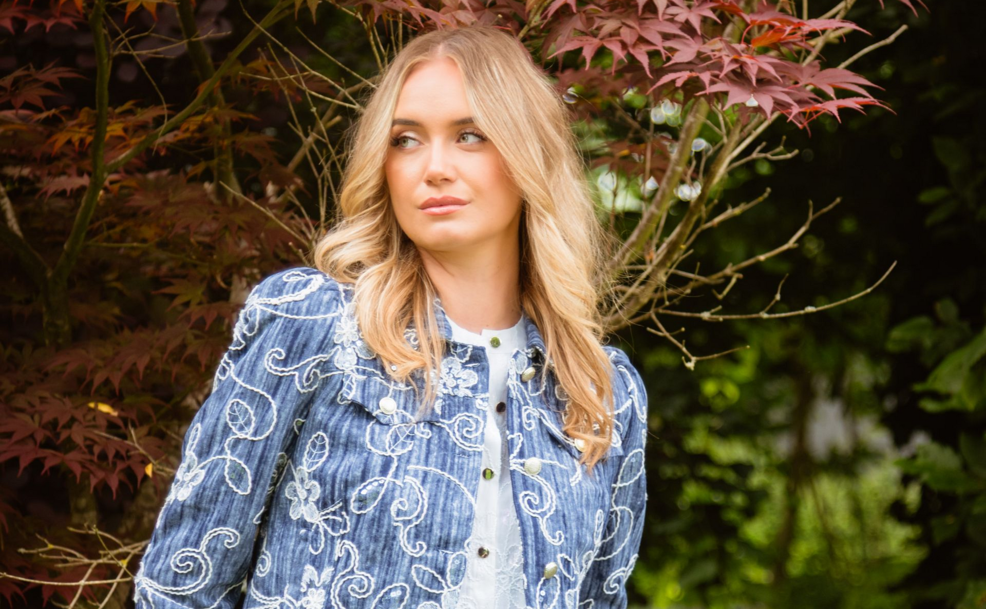 Blonde vrouw in een blauwe jas met patroon, buiten, kijkt weg van de camera, vlakbij een boom met rode bladeren.