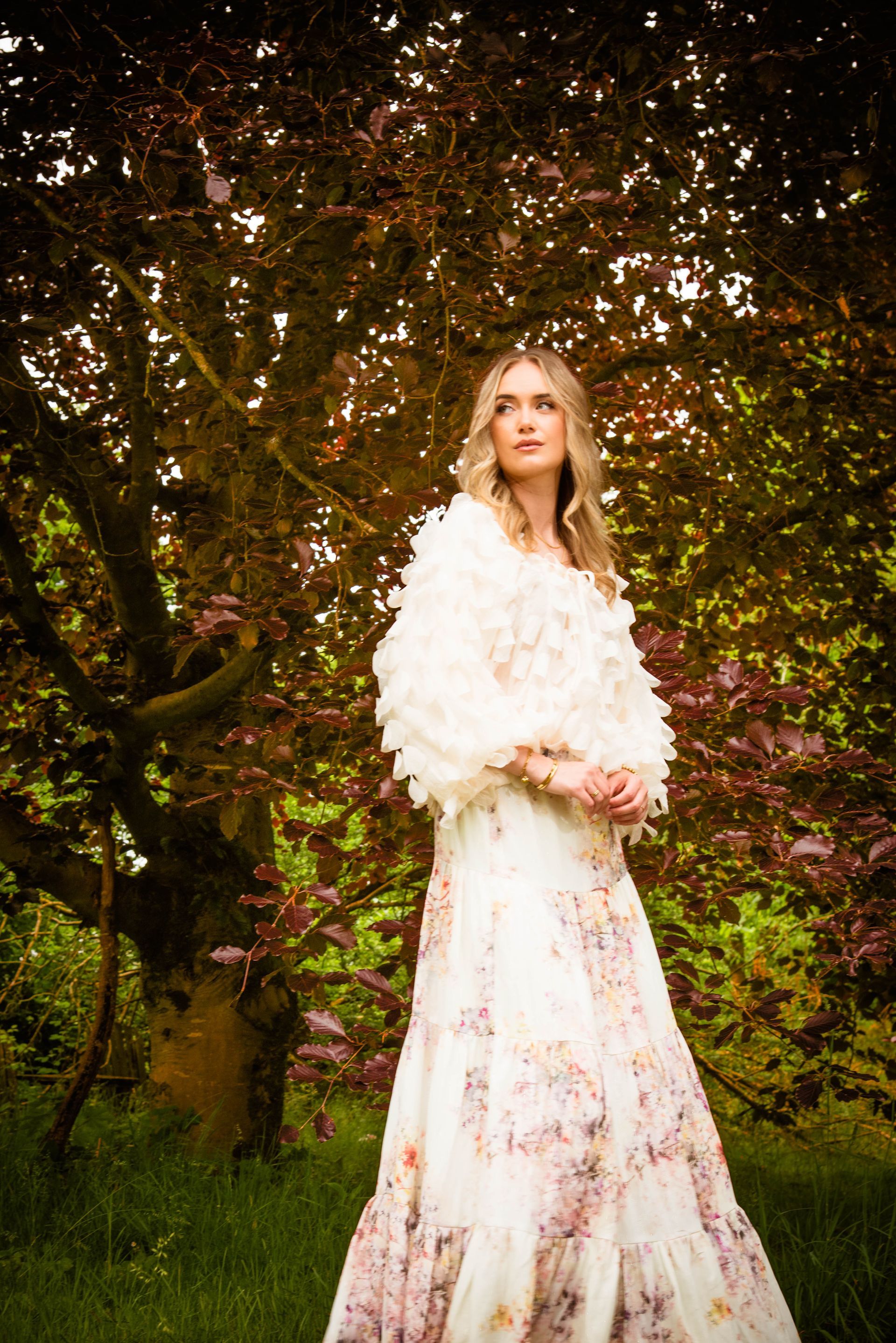 Een vrouw in een jurk met bloemenprint en een witte sjaal poseert onder een boom met roodachtige bladeren.