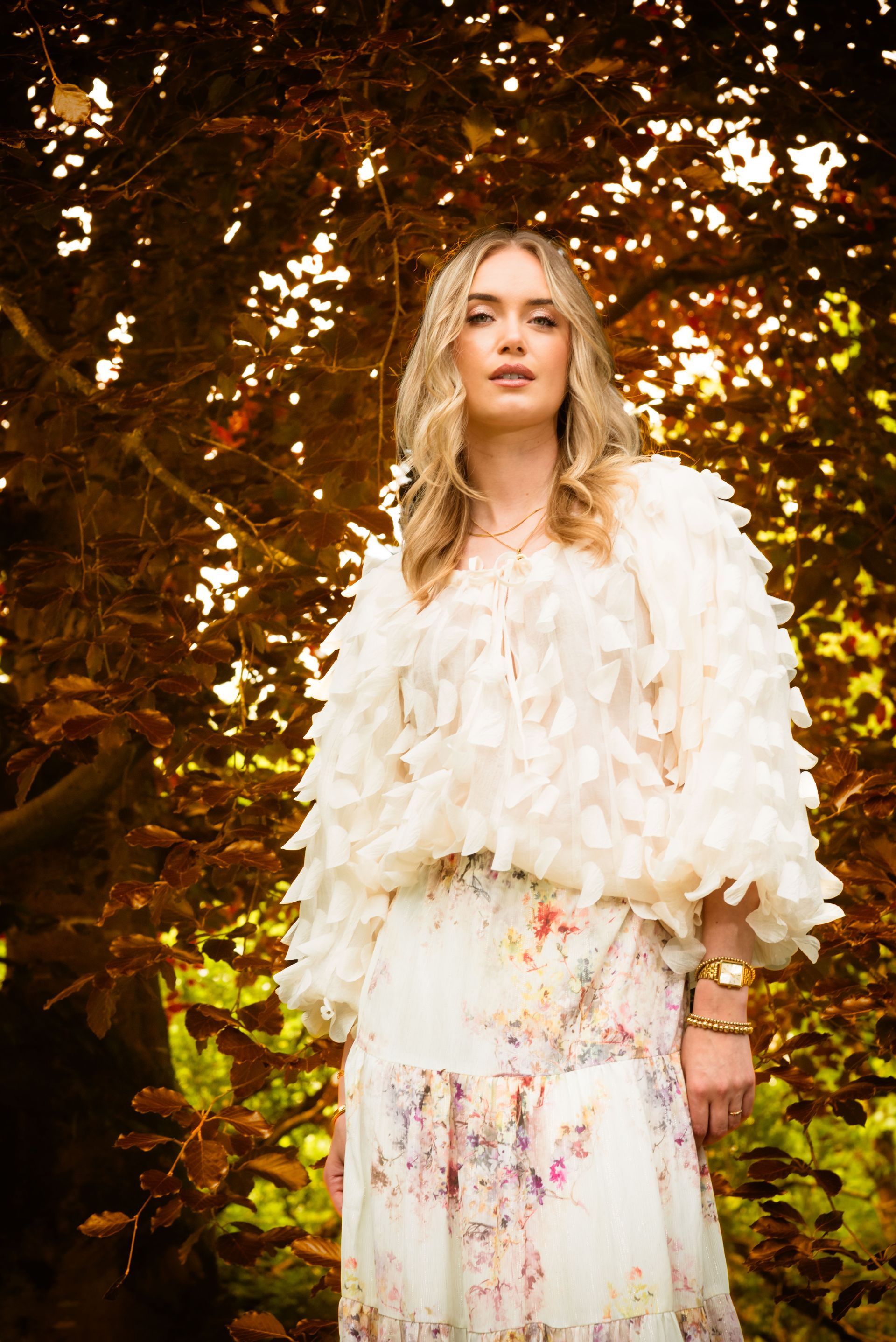 Vrouw in crèmekleurig jasje met franjes en bloemenrok, staand voor herfstbladeren.
