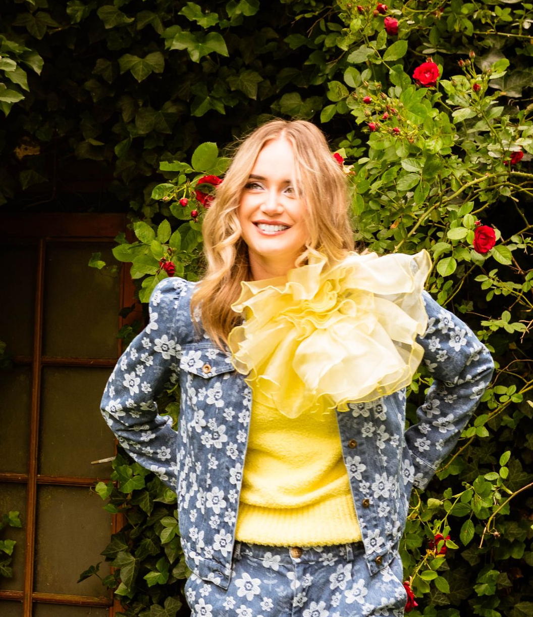 Een vrouw in een spijkerjasje met bloemenprint en een gele trui lacht voor een rozenstruik.